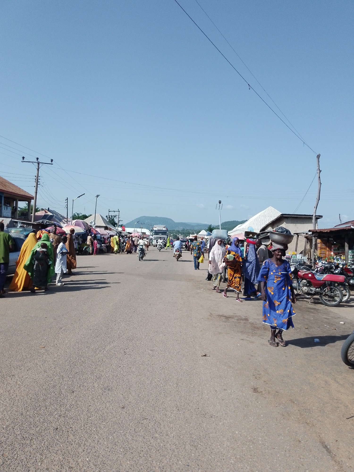 The image shows a bustling street scene in Serti, Nigeria. People are walking along a paved road lined with market stalls and shops. Several individuals are carrying goods on their heads, including a woman in a blue dress with yellow and orange patterns. There are motorcycles parked and being ridden on the road, and a large truck is visible in the distance. The sky is clear and blue, and the scene appears to be set during the daytime. The overall impression is one of a lively and active marketplace.