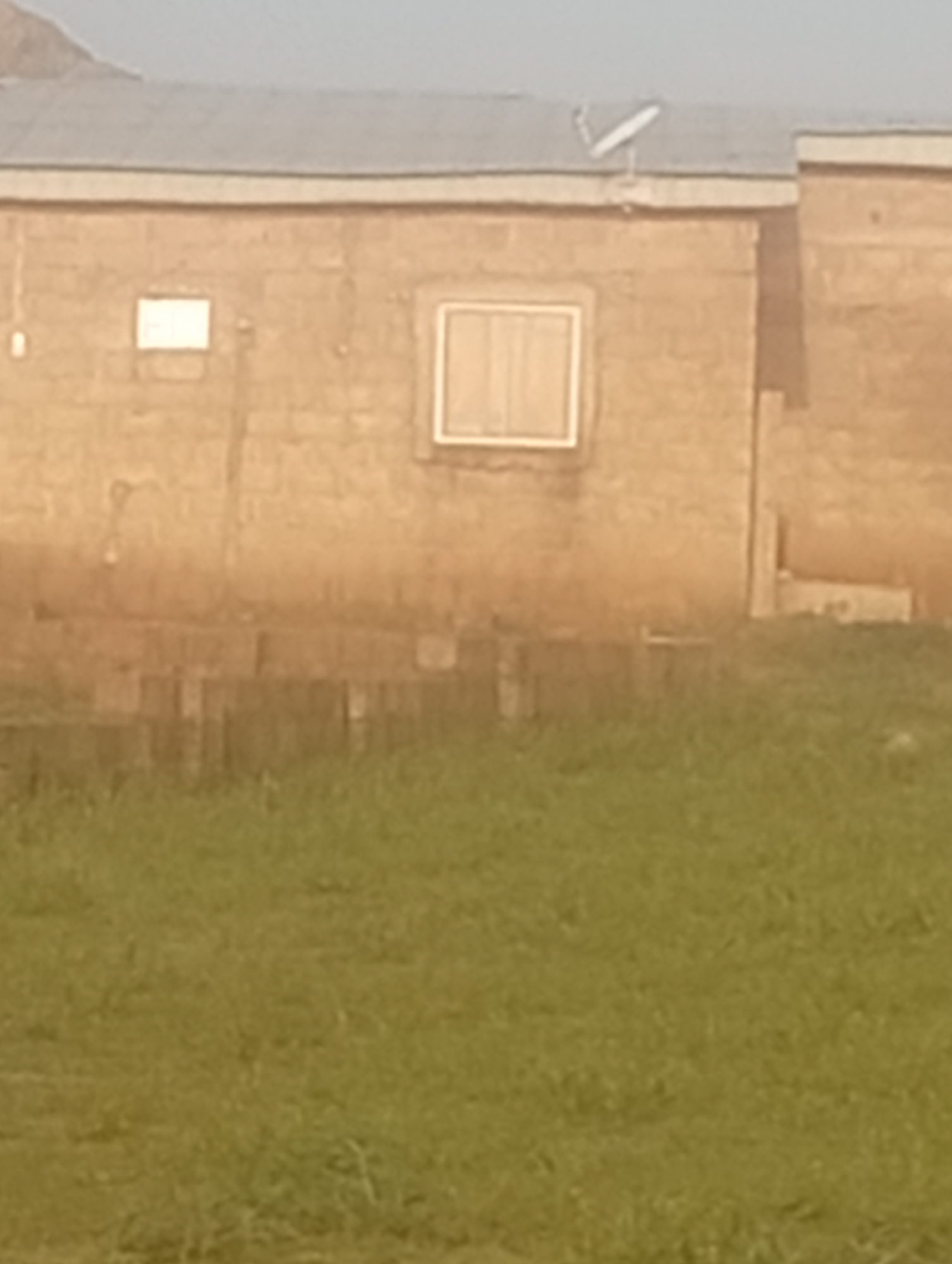 A medium shot captures a tan, brick-building structure with a grey corrugated metal roof in Katon Rikkos, Nigeria. The building has one visible window, a square pane with white trim and shutters, and a smaller rectangular opening to its left. A satellite dish is affixed to the roof on the right side of the structure. In front of the building, a patch of unkempt green grass and what appears to be stacked cinder blocks is visible. The overall lighting suggests daytime, with a slight haze obscuring distant details.
