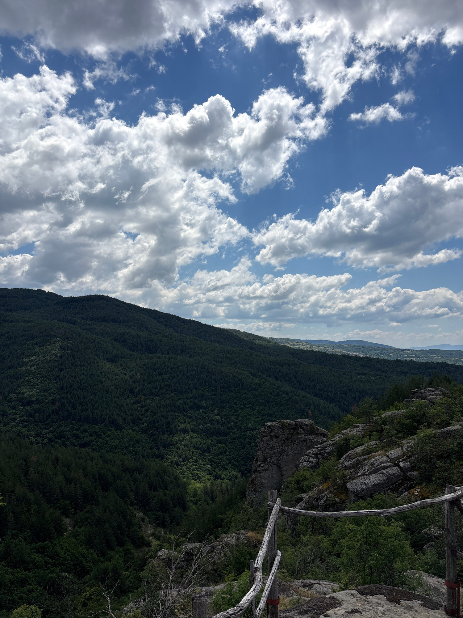 The image depicts a panoramic view from a rocky, elevated vantage point overlooking a lush, forested valley. The sky above is a vibrant blue, adorned with scattered, fluffy white clouds. The foreground features a rustic wooden railing, weathered and aged, with a few exposed rocks. Beyond the railing, the terrain drops into a dense, dark green forest that covers the slopes of a large hill or mountain. In the distance, the valley opens up to reveal more rolling hills and a distant settlement with scattered buildings, suggesting a rural area. The overall impression is one of natural beauty and tranquility. The location is identified as being in Dolno Drianovo, Bulgaria. There are no people or animals visible in the image.