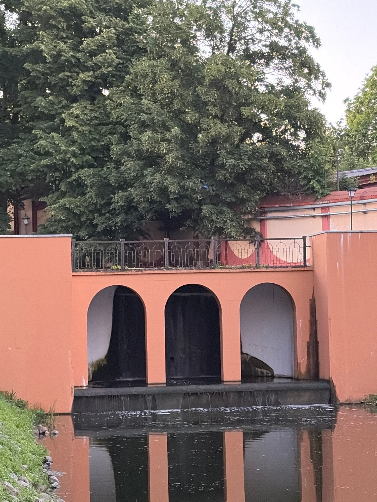An outdoor, medium-range, eye-level shot shows a structure with three arched openings built into an orange-painted wall. The middle and right openings are partially obscured by dark material, while the left opening is more clearly visible and appears to be a dark, recessed doorway. Water flows over a low barrier in front of the arches, creating a small waterfall. The water then collects in a body of water in the foreground, reflecting the arches and the orange wall. Above the arches, a black metal railing with decorative scrollwork runs along a raised walkway. Beyond the railing, a large, leafy green tree dominates the upper portion of the frame, with its branches reaching across the sky. In the background, sections of a building with red-tiled roofs and cream-colored walls are visible, along with a black lamppost. The scene is well-lit, suggesting it is daytime. There are no people visible in the image.