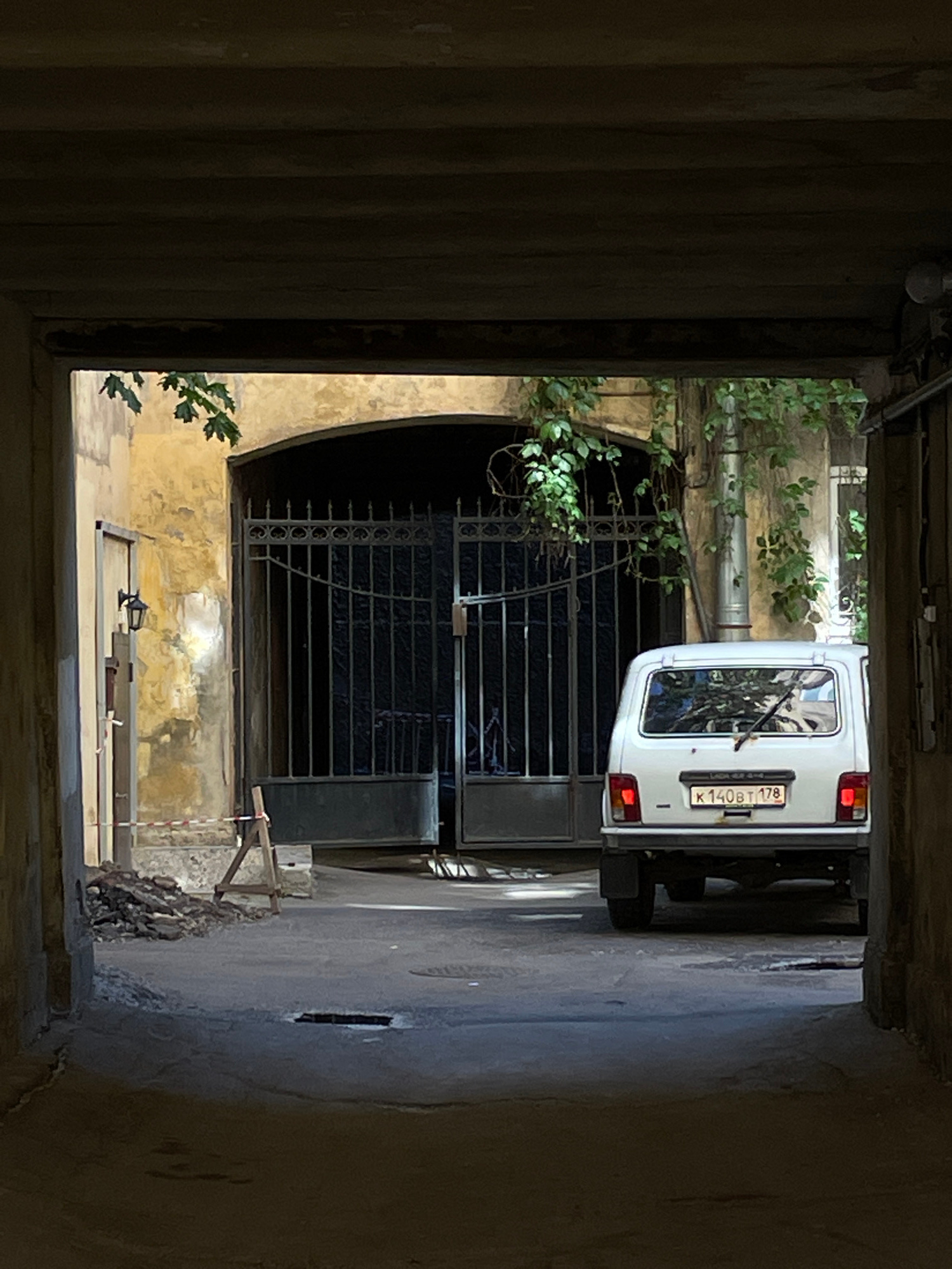 The image shows a view looking down a narrow, shadowed passageway in what appears to be an older urban setting. On the left side of the passageway, a ribbed, dark vertical structure dominates the foreground, possibly a corrugated metal wall or a similar architectural element. The passageway opens up into a brighter area where a white van is parked, its rear end facing the viewer. The van has a license plate that reads "K 140BT 178".
Behind the van, a large, ornate wrought-iron gate is visible, partially obscured by the van and by lush green foliage growing on the left side. The gate appears to lead to an inner courtyard or building. The walls of the passageway and the building beyond are weathered and textured, with peeling paint and visible brickwork in places, suggesting age.
In the foreground of the brighter area, beneath the gate and to the right of the foliage, there is a small, rough wooden structure that looks like scaffolding or a temporary support. Some debris and rubble are visible near the base of the wall on the right.
The lighting suggests it is daytime, but the shadows within the passageway create a sense of dimness and confinement. The overall impression is one of a slightly neglected but functional urban space, perhaps a service entrance or a private yard in an older part of Saint Petersburg, Russia. There are no people visible in the image.