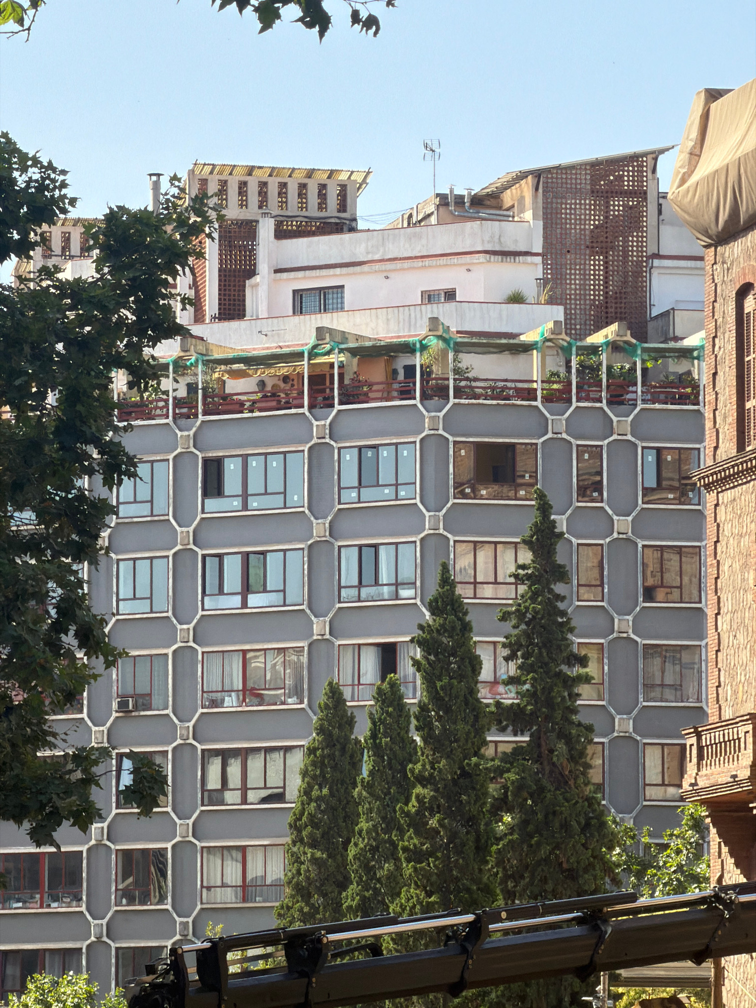 The image shows a multi-story building with a grey facade, featuring a distinctive geometric pattern of windows and balconies. The building is surrounded by trees, with some foliage obscuring parts of the structure. In the background, other buildings are visible, including one with a brick facade and intricate architectural details. The sky is clear and blue. The scene is located on Carrer de l'Eixample in Barcelona, Spain, as identified by its unique architectural style and urban setting. There are no people or specific actions depicted in the image, focusing solely on the architectural elements and their context within the urban environment.