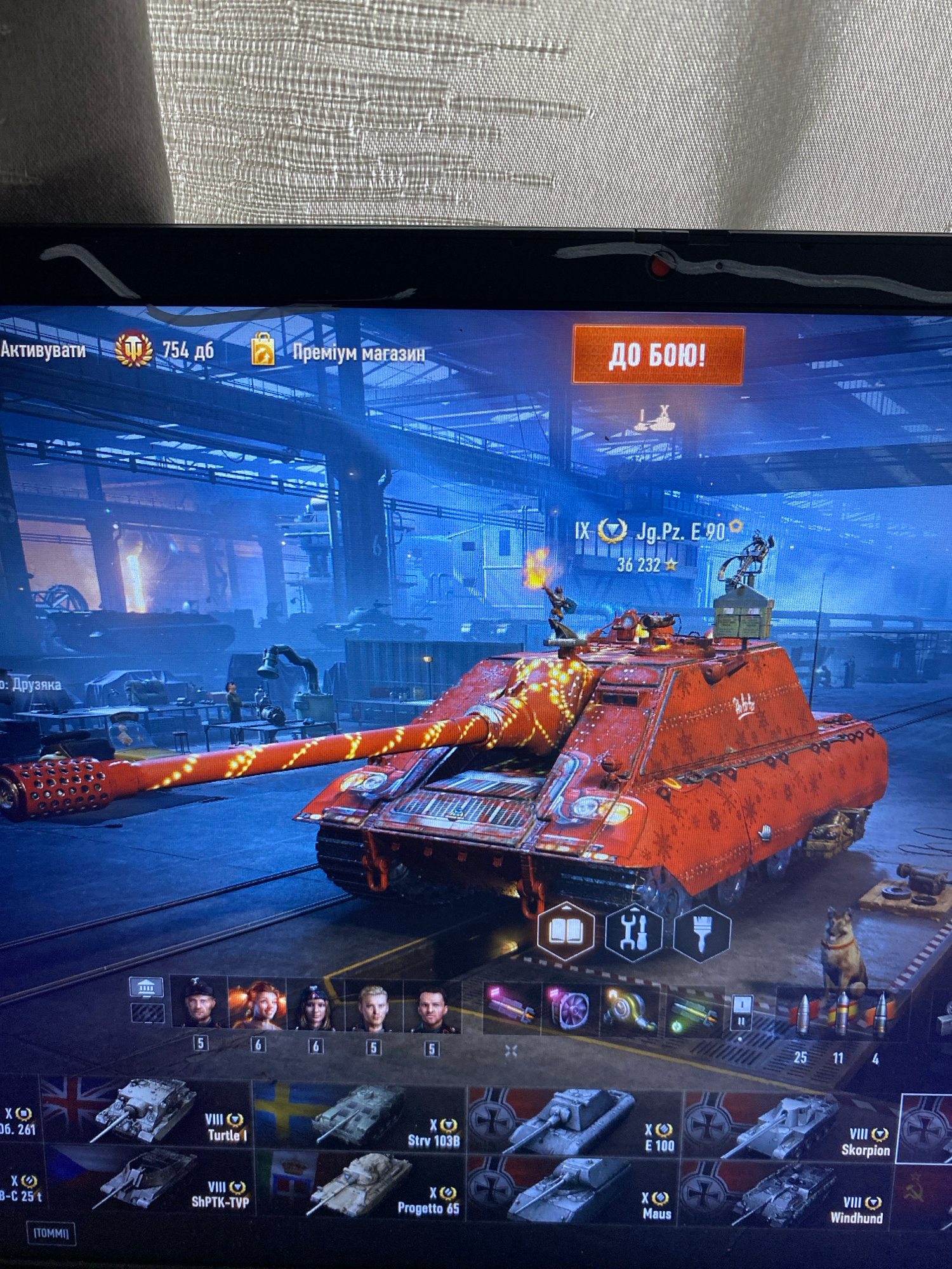 The image displays a computer screen showing a video game interface, specifically a vehicle combat game. The game is set in a virtual environment that appears to be a hangar or workshop, with a large, heavily armored red tank with golden accents prominently featured in the foreground. The tank has intricate detailing and appears to be customized with a unique paint job.
In the upper left corner of the screen, text is visible in Ukrainian, including "Активувати" (Activate) and "Преміум магазин" (Premium shop), along with a number indicating in-game currency or items. To the right, a large orange button reads "ДО БОЮ!" (To battle!). Above the tank, the designation "IX Jg.Pz. E. 90" is displayed, indicating the type and tier of the vehicle. Below this, a numerical value of "36 232" is shown, likely representing experience points or a similar game metric.
The bottom portion of the screen shows a selection of other vehicles, each represented by an icon and a brief description, suggesting a player's garage or inventory. Some of the vehicle designations visible include "VIII Turtle I", "X Sterv 103B", "VIII ShPTK-TVP", and "X Progetto 65".
The environment depicted within the game is dimly lit, with blue and orange lights casting an atmospheric glow. There are other tanks visible in the background, along with industrial-looking structures and equipment. A dog is also visible in the lower right corner of the game scene, seemingly part of the virtual environment.
There is no visible indication or context within the image that links it to the city of Szczecin, Poland. The scene is entirely contained within the computer game's virtual world.