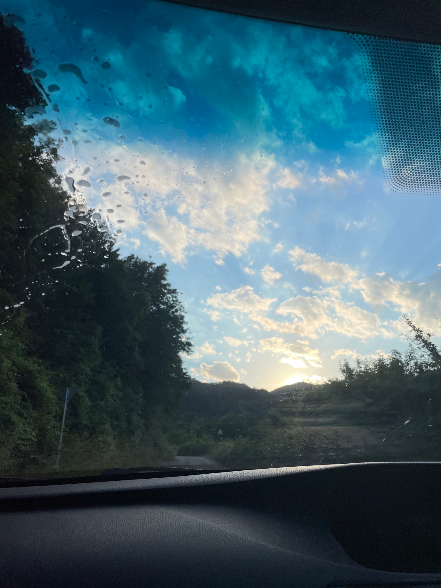 The image captures a view from inside a vehicle, looking through the windshield. The windshield has several water droplets and streaks on it, obscuring some of the view. The sky is partly cloudy with the sun setting behind hills. The landscape outside appears to be rural, with dense green foliage on either side of a winding road. The road itself is narrow and appears to be paved. There are no people or animals visible in the image. The interior of the car is visible in the foreground, specifically the dashboard, which is dark and textured. The scene suggests a drive through a scenic, possibly mountainous area. The specific location in the city of Zlatograd, Bulgaria is not identifiable from the visual information in the image.