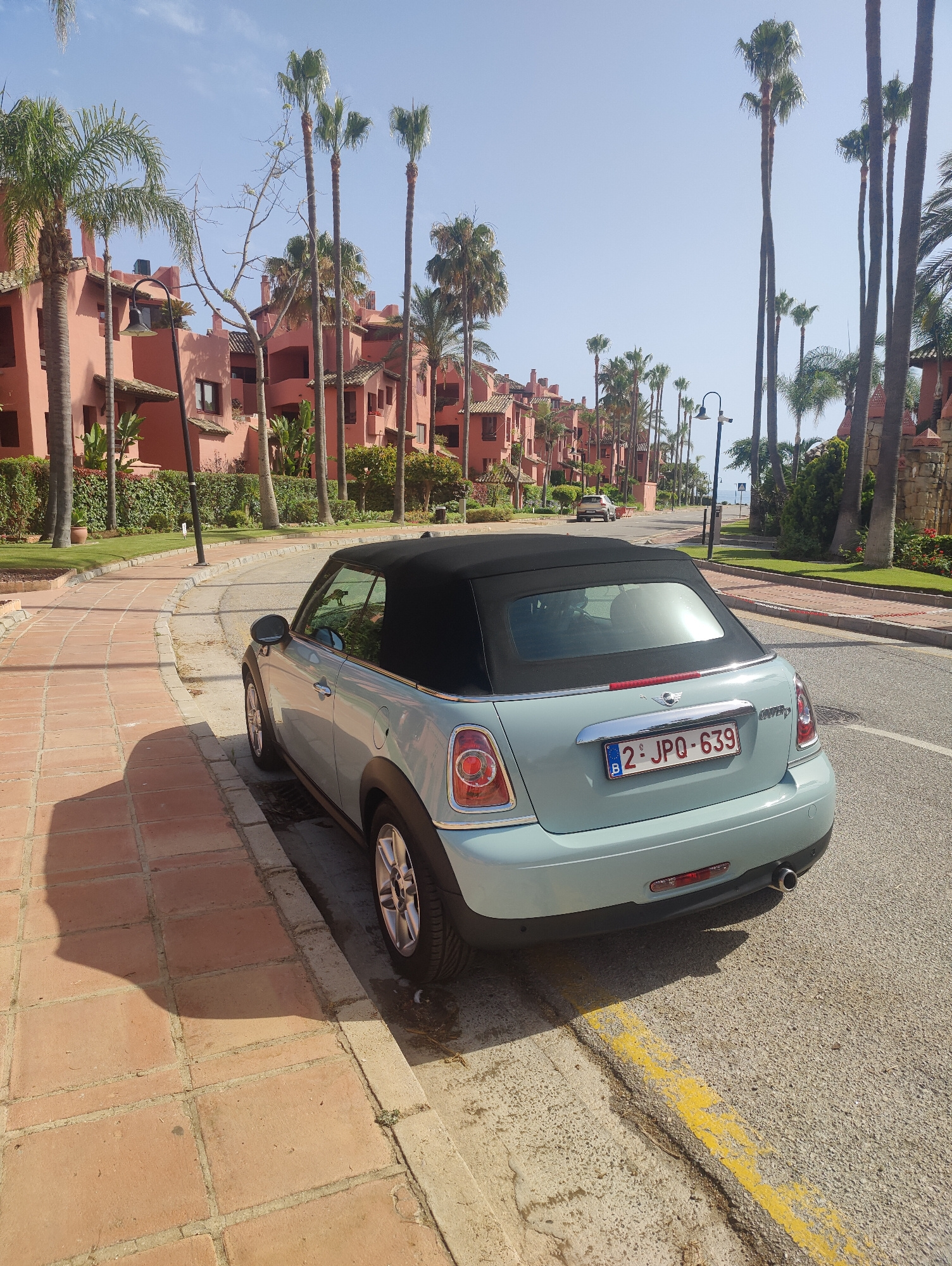 A light blue convertible Mini Cooper is parked on the side of a paved road, with its black convertible top up. The car is facing away from the camera, with its rear visible. The license plate reads "B 2-JPQ-639".
The car is parked on the edge of a sidewalk made of reddish-brown tiles. The sidewalk is bordered by a curb and then the asphalt road. To the left of the sidewalk, there is a manicured green lawn. Beyond the lawn, there are several rows of pink buildings with red-tiled roofs, characteristic of Mediterranean architecture. Tall palm trees are scattered throughout the scene, some lining the road and others in front of the buildings. The sky is clear and blue, suggesting a sunny day. The sun casts long shadows from the palm trees and the car onto the sidewalk and road. In the background, a hint of the sea can be seen between the buildings and palm trees, with a beach umbrella on the shore. The overall impression is a sunny day in a resort or residential area, likely in a warm climate. There are no people visible in the image. The time of day appears to be late morning or early afternoon, given the angle of the shadows.