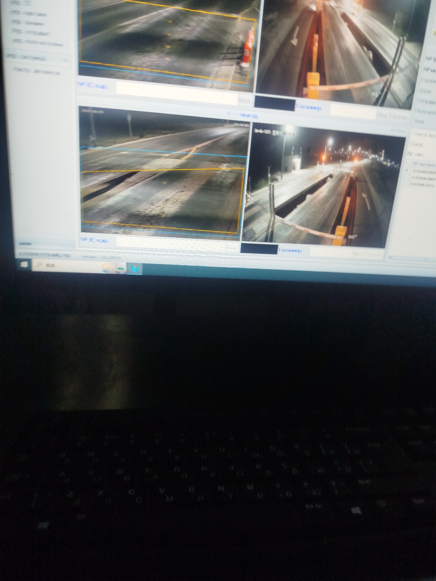 The image shows a computer screen displaying a CCTV surveillance system. The screen is divided into multiple frames, each showing a view of a road, likely at an inspection or checkpoint. One frame displays a close-up of traffic cones. Another frame shows a wider view of a road with barriers and what appears to be a toll booth or inspection station. Text labels in Russian, such as "Госномер" (license plate) and "Выезд" (exit), are visible. The lower part of the image shows a dimly lit keyboard of a laptop, suggesting the screen is part of a laptop setup. The location "Түндүк айланып өтүүчү жол 84 га, Kyrgyzstan" is a specific address in Kyrgyzstan.