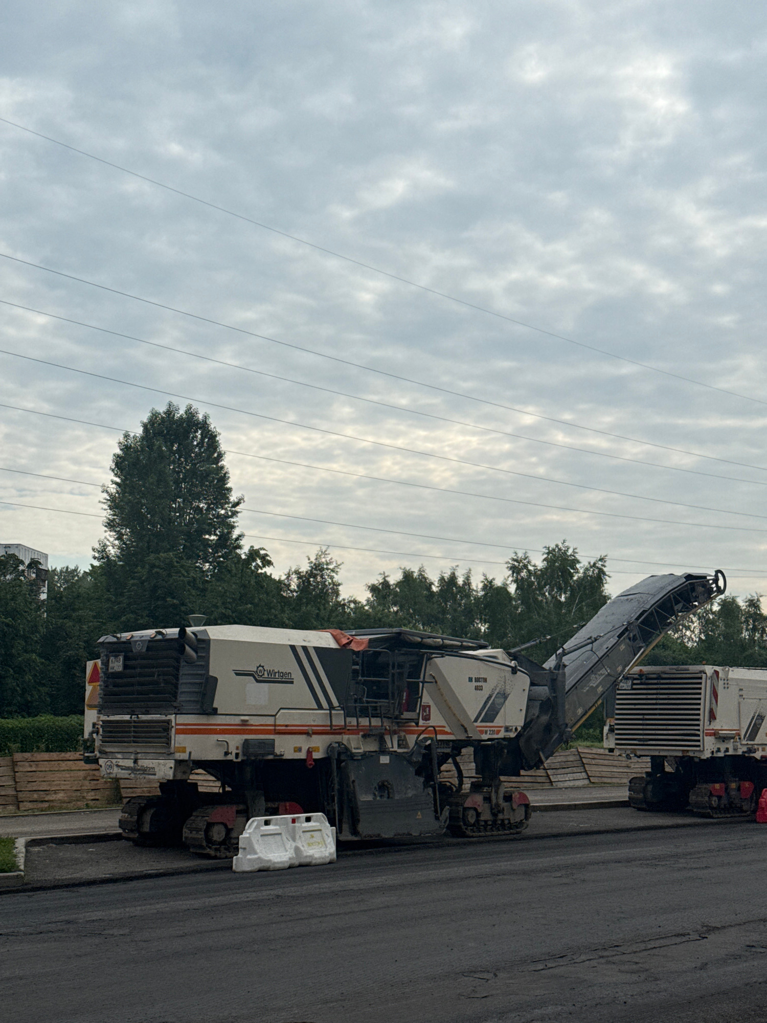 The image depicts a road milling machine, likely a Wirtgen brand, operating on asphalt. The machine is white with black and orange accents and has caterpillar tracks. A conveyor belt extends from the machine, indicating it is actively removing material from the road surface. Two white plastic barriers are placed on the asphalt near the machine. In the background, a line of trees separates the road from a building and power lines are visible against an overcast sky. The scene suggests ongoing road construction or maintenance work. While the image does not explicitly show the city of Moscow, Russia, road milling machines of this type are commonly used in urban infrastructure projects.