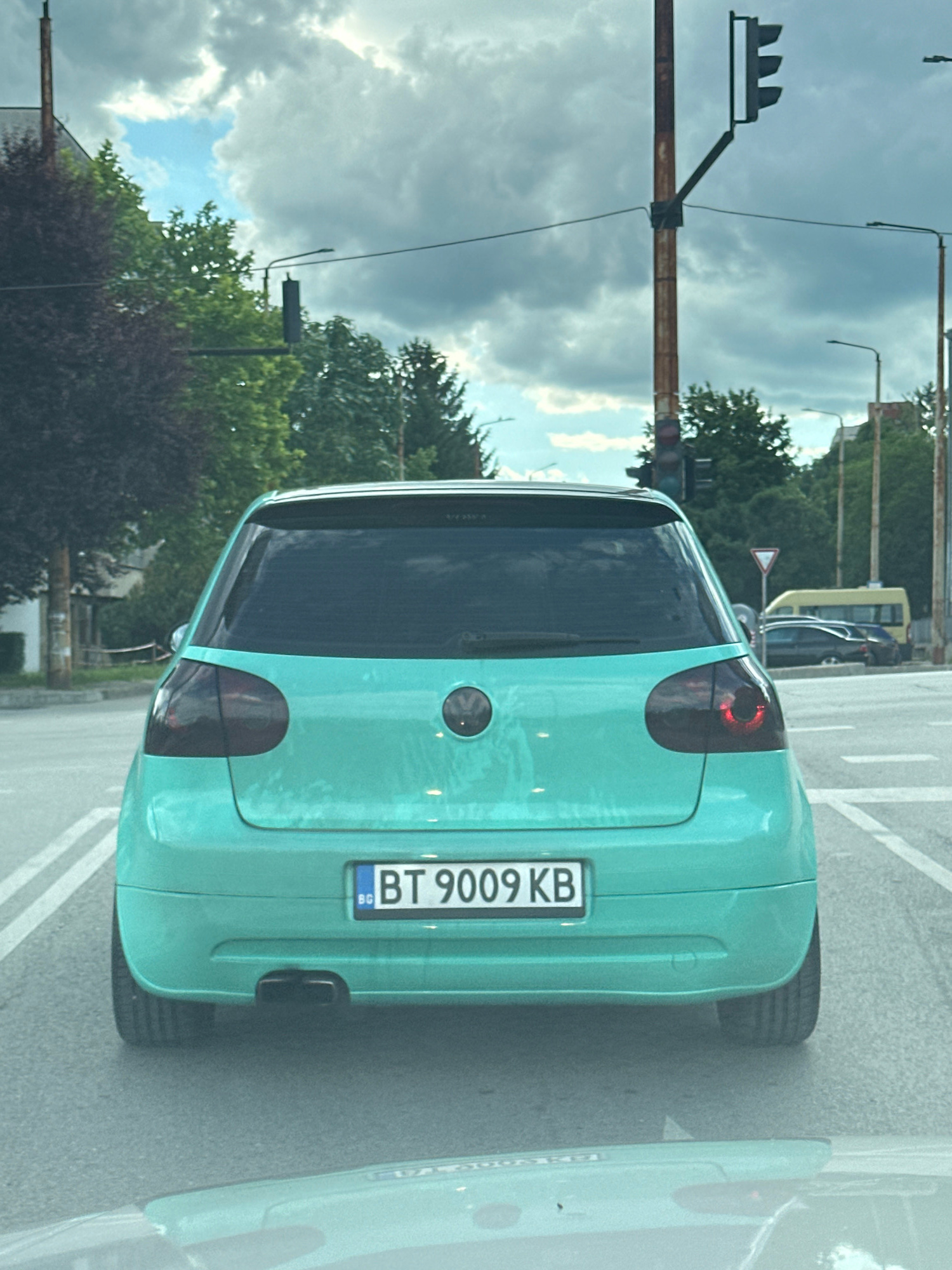 A turquoise Volkswagen Golf is viewed from the rear, stopped at a traffic light in Veliko Tarnovo, Bulgaria. The car has tinted taillights and a license plate that reads "BT 9009 KB". In the background, there are trees, buildings, and traffic lights. The sky is cloudy.