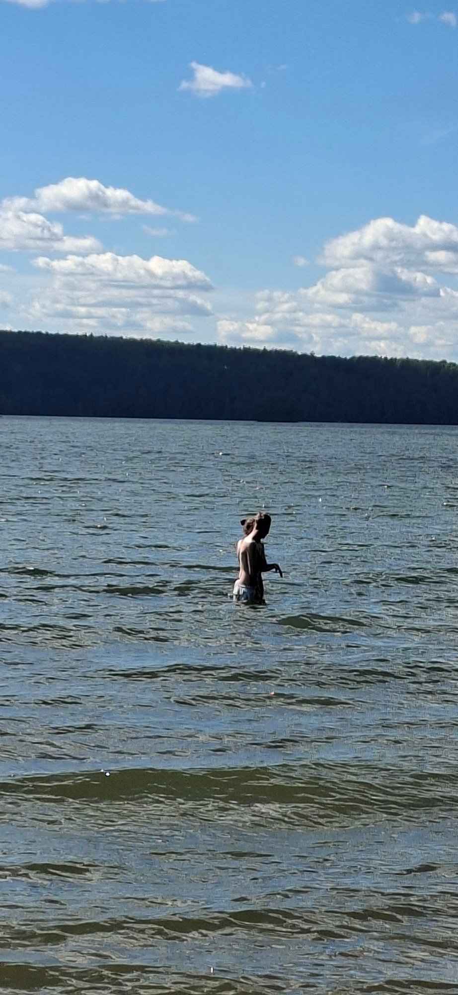 This is a vertically oriented, wide-angle photograph taken outdoors during the daytime. The scene depicts a person swimming in a body of water, likely a lake, with a tree-covered shoreline in the background. The water is calm, with gentle ripples reflecting the sunlight. The sky above is a clear blue, with scattered white, fluffy clouds. The person appears to be a man, wearing shorts and is partially submerged in the water, with his arms raised and head tilted back, possibly in a relaxed or playful pose. The image is well-lit, suggesting it was taken on a sunny day. The overall mood is serene and peaceful, capturing a moment of enjoyment in nature. There is no visible text in the image. The location is Lahti, Finland.
