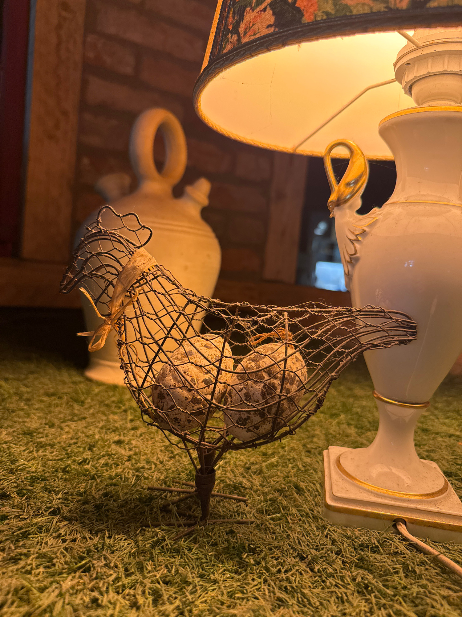 This media file features a close-up, vertically oriented shot of decorative objects arranged on a faux grass surface. The primary subjects are two cream-colored ceramic vases, one taller with gold trim and ornate detailing, and another shorter, jug-like vase with a loop handle.
Dominating the foreground is a wire cage containing what appear to be several speckled eggs. The cage itself is crudely formed, with a small strip of light-colored material tied around its opening. The eggs are clustered together within the cage.
Behind the wire cage, and partially obscured, is a lamp with a patterned lampshade, emitting a warm, yellow light that illuminates the scene. In the background, the base of the taller vase is visible, resting on the textured green surface. The overall impression is of a still life arrangement, possibly a decorative display for a garden or indoor setting.
The lighting suggests it might be evening or indoors, with artificial light providing illumination. There are no people visible, and the focus is entirely on the inanimate objects. The textures of the ceramic, wire, eggs, and faux grass are all discernible. There is no visible text in the image.