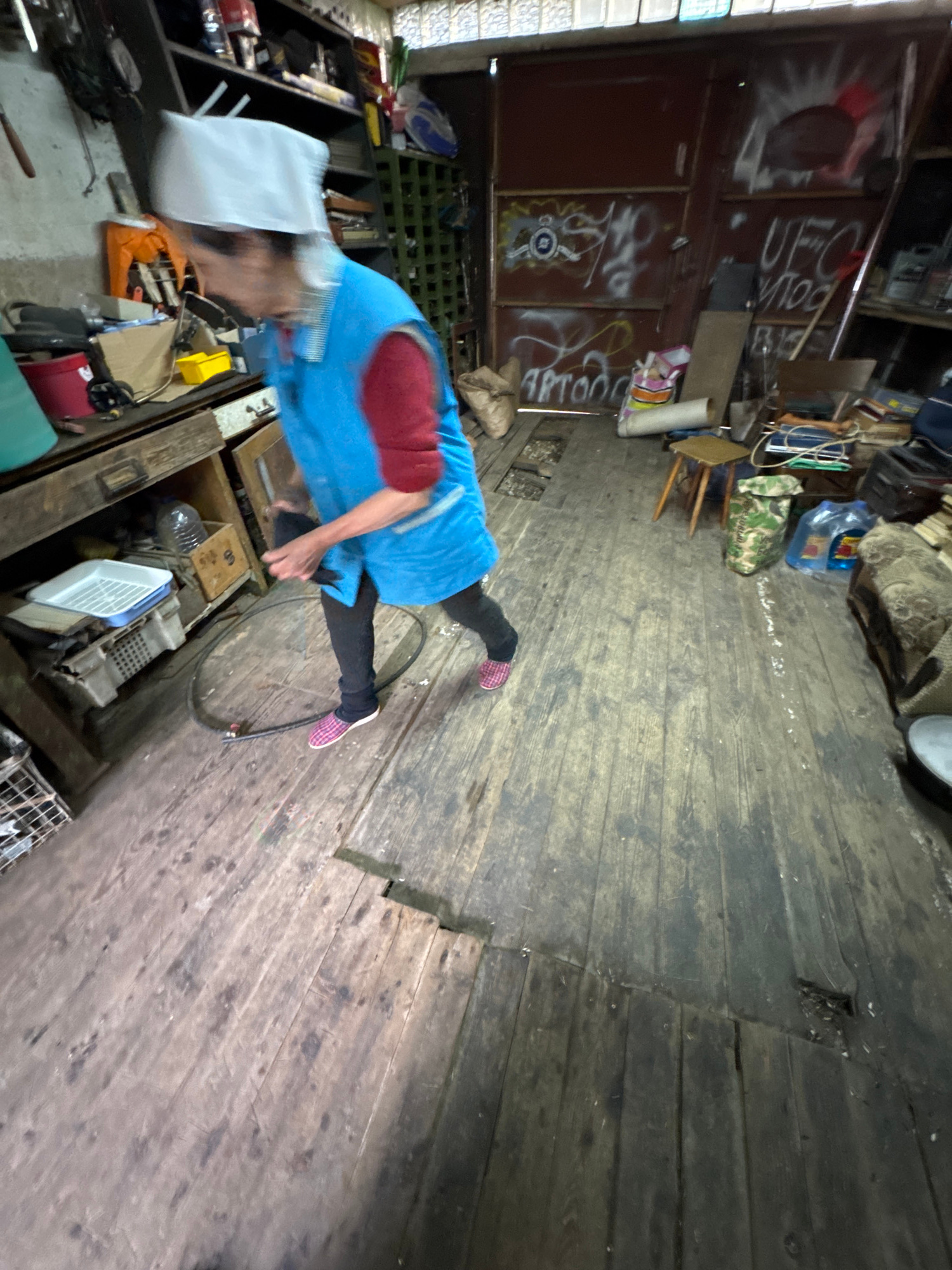 A medium shot, captured from a slightly elevated angle, shows an elderly woman wearing a white head covering and a blue apron over a red long-sleeved shirt and dark pants. She is walking on a weathered wooden floor. Her right hand is extended, and she appears to be holding a black hose or cable. The room is cluttered with various items, suggesting a workshop or storage area. To the left, shelves are filled with boxes and containers. In the background, a large metallic door is covered in graffiti. The floor is made of worn wooden planks, some of which are broken or have gaps, revealing dirt and debris below. The overall environment appears unkempt and industrial. The location is specified as Naro-Fominsk, Russia.