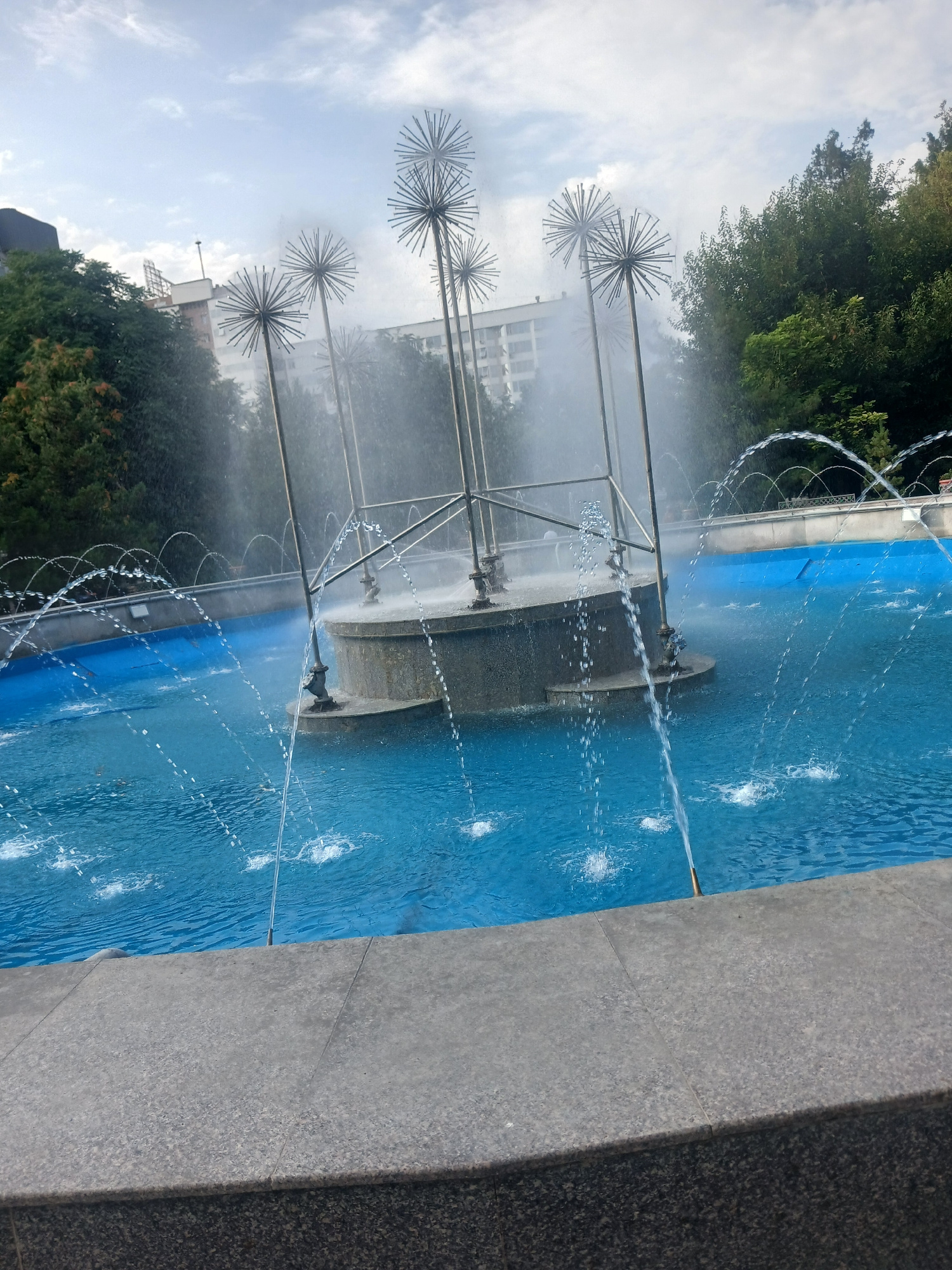 The image depicts an outdoor fountain in Shymkent, Kazakhstan. The fountain features a central, circular structure from which water jets emanate upwards, creating a mist. Surrounding the central structure, there are several tall, thin poles topped with spherical, spiky metal sculptures resembling dandelions. The fountain is filled with bright blue water, and smaller water jets shoot up from various points within the pool. In the background, there are trees and modern buildings, suggesting an urban park or public square setting. The sky is partly cloudy. The foreground shows a grey, textured stone ledge. No people are visible in the image.