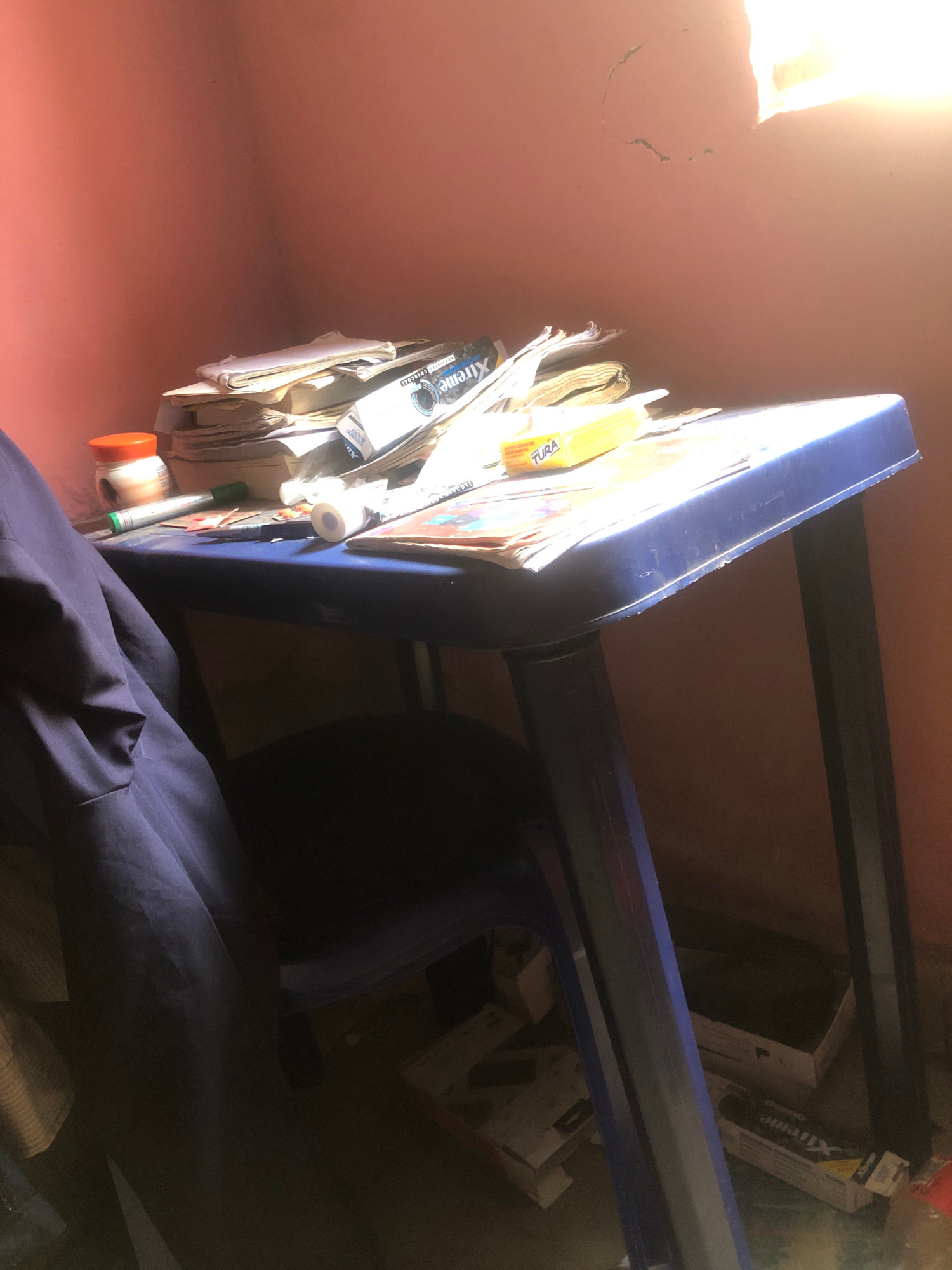 A low-angle, vertically oriented shot captures a shelf unit overflowing with various items, including what appears to be papers, books, and packaged goods. The shelf unit, a dark blue color, is positioned against a pale pink wall. A bright light source, possibly sunlight, illuminates the upper left portion of the image, casting a warm glow and creating a hazy effect.
On the shelf, a stack of papers or books is visible, some with what looks like yellow packaging or labels. One label clearly shows the word "TURA." Further down, another item has the brand name "Xtreme" printed in white text on a blue background. Several cylindrical objects are also present, one with an orange cap and another that appears to be a roll of tape or a small bottle with a white cap. A green marker pen is also visible among the clutter.
Beyond the shelf, the scene recedes into shadow, revealing more of the shelf unit's structure and what might be boxes or other items stored on lower levels. The lighting suggests it is daytime, with the bright light source indicating either morning or afternoon sun. The setting appears to be indoors, possibly a room or storage area. The presence of what looks like personal belongings and office or school supplies suggests a domestic or study environment. The image is rich with detail due to the close-up perspective, highlighting the textures and colors of the objects on the shelf.