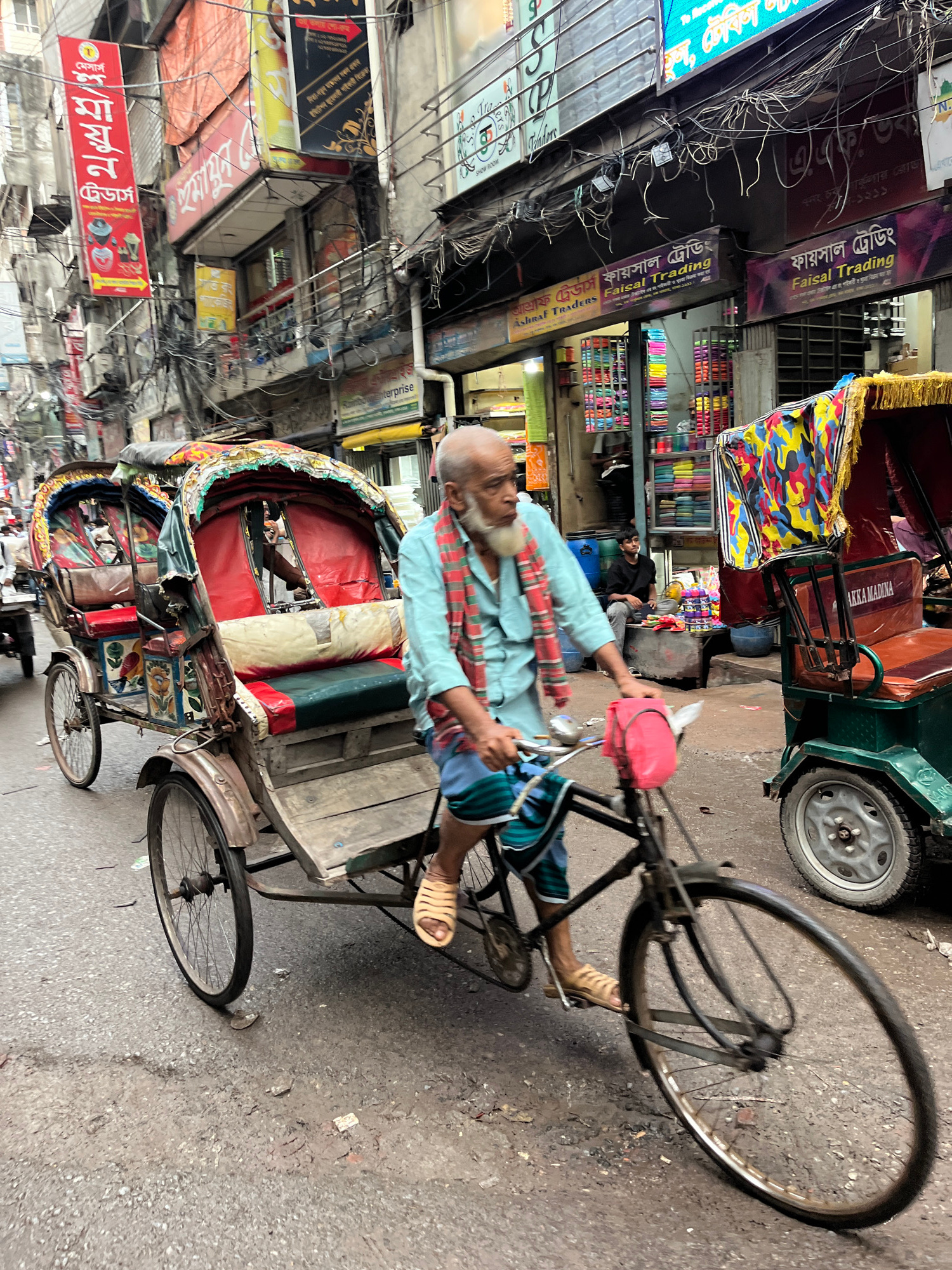The image depicts a busy street scene in Dhaka, Bangladesh, likely a market area given the numerous shops and signage. In the foreground, an elderly man with a white beard, wearing a light blue shirt and a red and blue checkered scarf, pedals a cycle rickshaw loaded with passengers. The rickshaw has a vibrant, colorful canopy and is decorated with floral motifs. The man is wearing tan sandals. Behind him, another similar rickshaw is partially visible. The street is paved with asphalt and appears somewhat dirty. The buildings lining the street are multi-story, with visible electrical wires and numerous signs in Bengali script advertising various businesses. One prominent sign says "Faisal Trading" in both Bengali and English. A young man in a black shirt is seated on a ledge in the background, near a shop displaying stacks of colorful textiles. The overall atmosphere is one of bustling urban life.