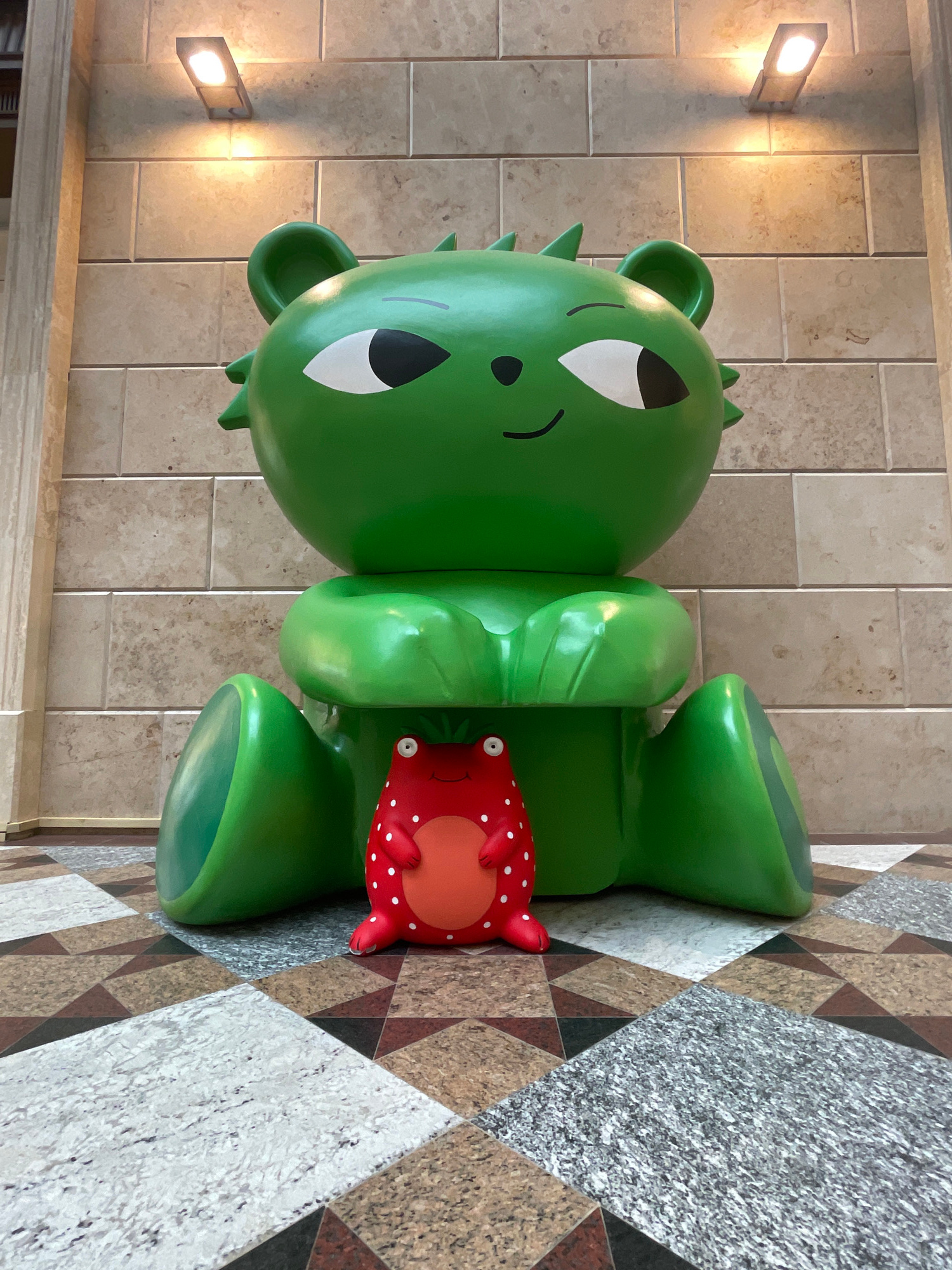 An overhead view from a low angle captures a large, bright green, cartoon-like bear sculpture sitting on a patterned floor. The sculpture has large, white-outlined eyes with black pupils, a small black mouth, and pointed ears. Its arms are bent, resting in its lap, and its legs are splayed outward. In front of the green bear, a smaller, red, cartoon-like strawberry figure sits on the floor. This figure has white spots, round eyes, and a small smile. The floor is a mosaic of geometric shapes in various earthy tones and grays. Behind the sculptures, a wall of light beige, rectangular tiles is visible, with two rectangular light fixtures mounted on it, casting a warm glow. The overall scene appears to be indoors.