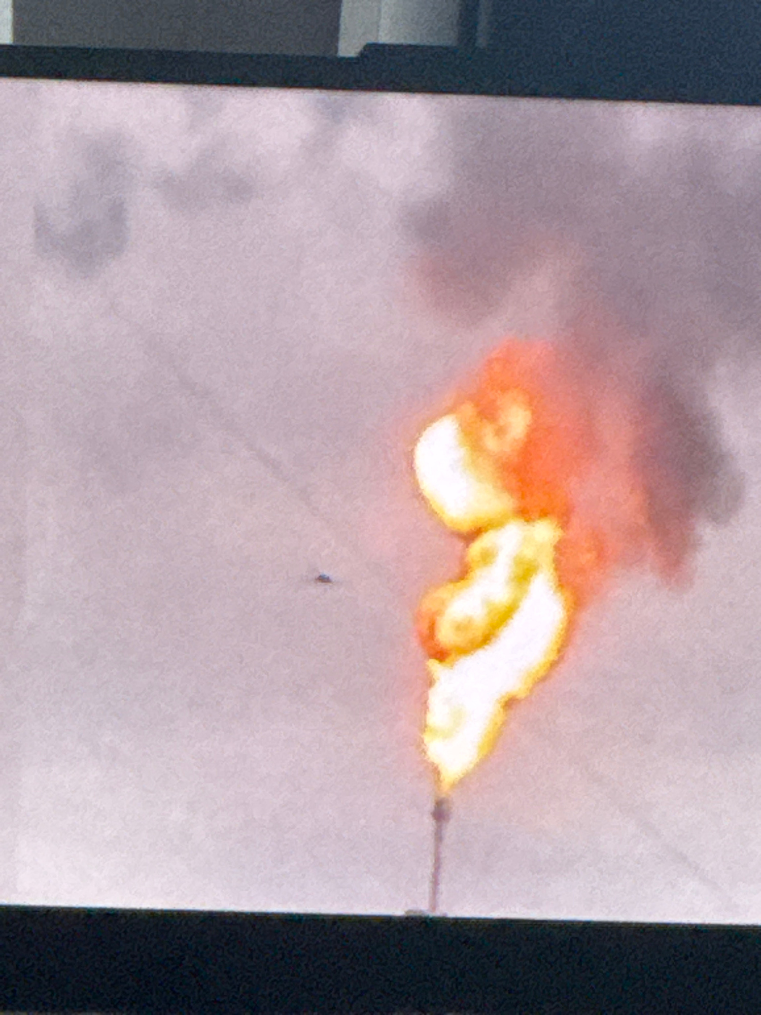 The image displays a flare stack emitting flames and smoke against a hazy, light-colored sky. The flare stack is a vertical structure with a flame at its apex. The flames are bright orange and yellow, and smoke, appearing as a grayish-purple cloud, billows upwards and to the right. The background is a uniformly textured, light gray or pale purple, suggesting a cloudy or overcast sky. In the lower portion of the image, a dark, horizontal line runs across the frame, likely the edge of a screen or display. There is a small, dark object with a somewhat triangular or wedge shape visible in the sky to the left of the flame, which could be a distant bird or an aircraft. The scene appears to be an industrial setting, typical of oil and gas facilities. There is no specific indication of the city of Tyumen, Russia, within the image itself.