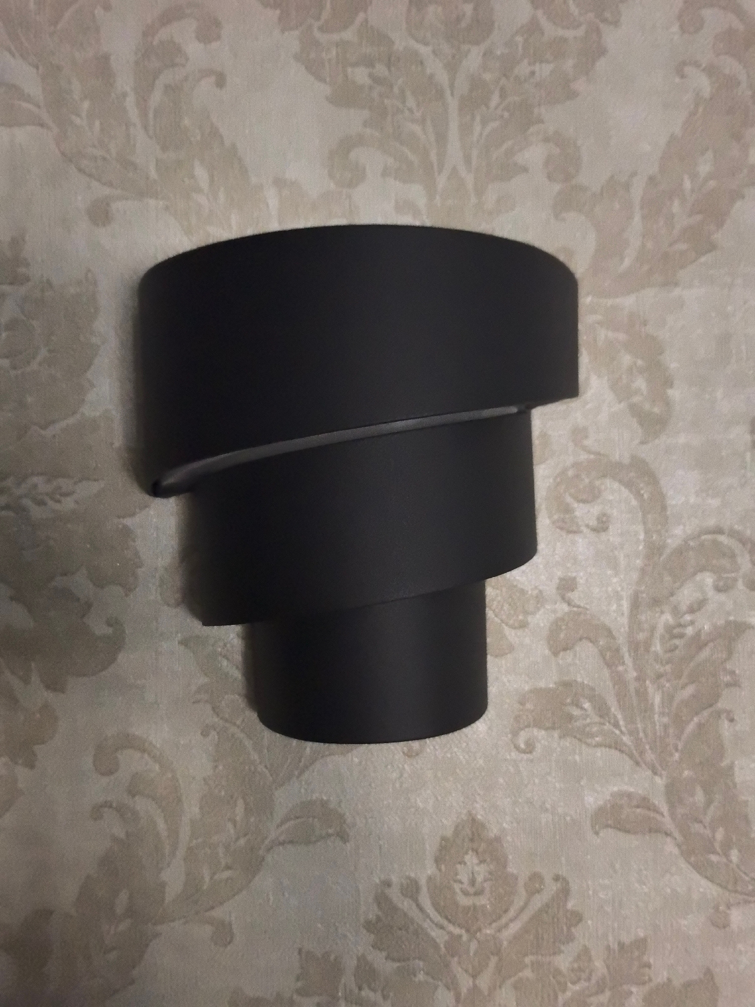 A black, modern, cylindrical sconce is mounted on a beige, patterned wallpaper. The sconce has three overlapping, curved segments that create a layered effect. The wallpaper features a light beige damask pattern on a slightly darker beige background. The image is a close-up, head-on view of the sconce. There are no people or other objects visible in the frame. The lighting appears to be from an interior source, casting a soft shadow on the wallpaper behind the sconce. The setting is likely an indoor residential or commercial space in Petropavlovsk-Kamchatsky, Russia.