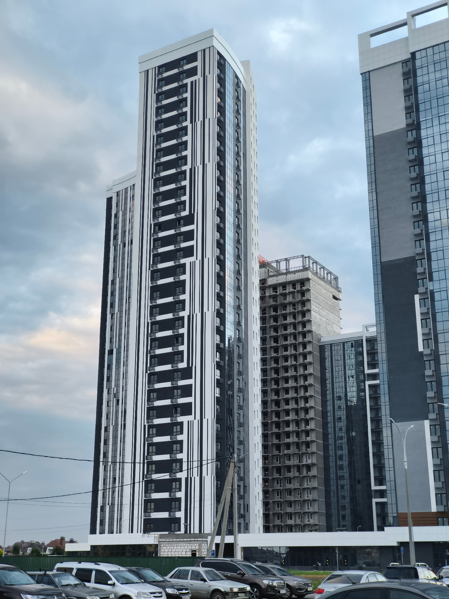 The image captures a view of a modern residential complex under construction and newly built high-rise buildings in Naberezhnye Chelny, Russia, under an overcast sky. The foreground shows a row of parked cars, including a dark SUV, several silver sedans, and other vehicles. A green fence separates the parking area from a grassy strip.
Dominating the mid-ground and background are several tall buildings. The most prominent is a striking black and white high-rise with a geometric facade, featuring vertical black stripes against a white background, interspersed with numerous dark-paned windows and small balconies. To its right, another skyscraper with a sleek, modern design is visible, primarily clad in reflective glass. Behind these, a multi-story building is clearly in the construction phase, with its concrete frame exposed and numerous open window frames. Another building with a glass facade is partially visible between the two main high-rises.
Utility poles and power lines are present in the scene, indicating urban infrastructure. In the distance, a few lower-rise buildings with sloped roofs can be seen, suggesting a mix of older and newer developments in the area. The overall atmosphere is one of urban development and ongoing construction.