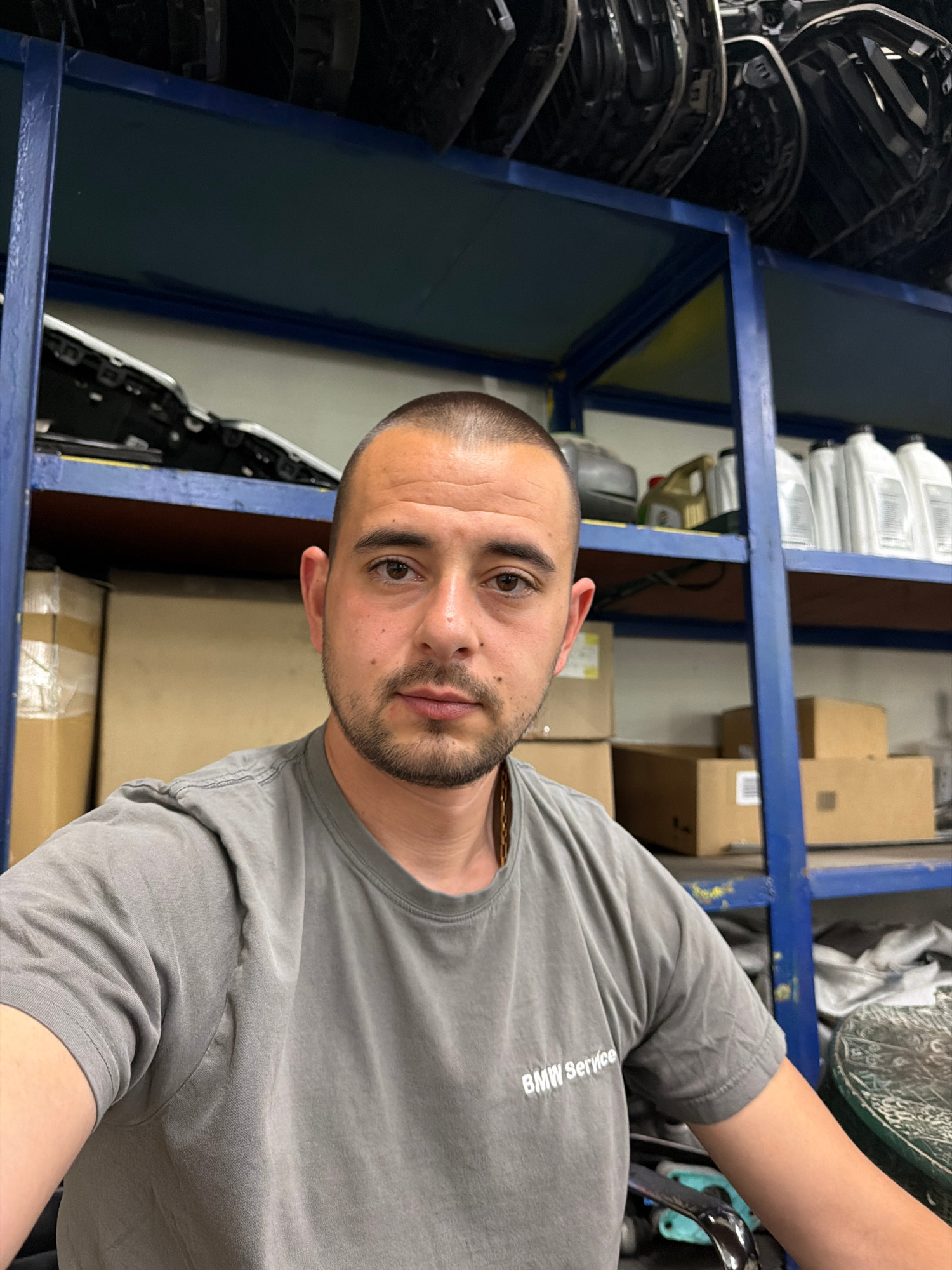 The image shows a medium shot of a man wearing a grey t-shirt with "BMW Service" printed on the left chest. He has short hair and a beard, and he is looking directly at the camera. The man is in what appears to be a garage or workshop, with shelves in the background holding various car parts and bottles of fluid. The shelves are blue metal, and some of the parts are car headlights. Boxes are stacked on shelves and on the floor. The lighting suggests an indoor environment, possibly illuminated by fluorescent lights. The overall impression is one of a professional automotive setting. The location is identified as Burgas, Bulgaria.