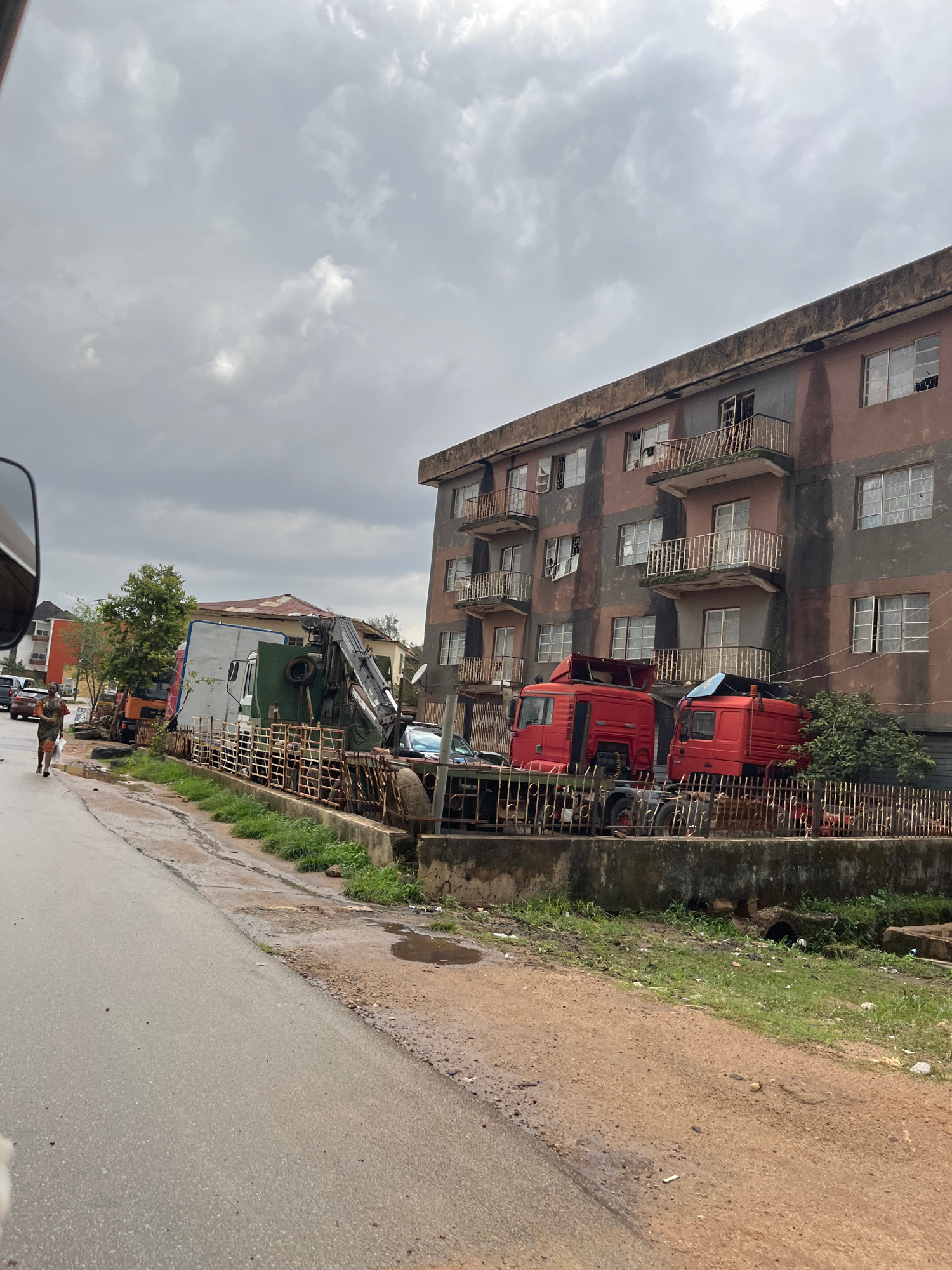 The image depicts a street scene in Jos, Nigeria, under an overcast sky, suggesting a cloudy or potentially rainy day. The perspective is from a vehicle, looking down a wet, paved road. To the left of the road, a multi-story building with a weathered, brownish facade stands. The building has numerous windows, many of which appear to be broken or boarded up. Balconies with rusted railings are visible on several floors.
Adjacent to the building and separated by a metal fence, several large red trucks are parked. One truck is positioned behind another, its cab clearly visible. Further down the street, a larger vehicle with a crane is parked, possibly a construction or utility vehicle. The area appears to be a mix of residential and commercial, with glimpses of other buildings and trees in the background.
The road surface shows signs of recent rain, with puddles and wet patches. The ground alongside the road is covered in patchy green grass and some debris. A person is walking on the right side of the road, wearing an orange top and dark trousers. The overall impression is one of a somewhat neglected urban environment. No visible text is present in the image.