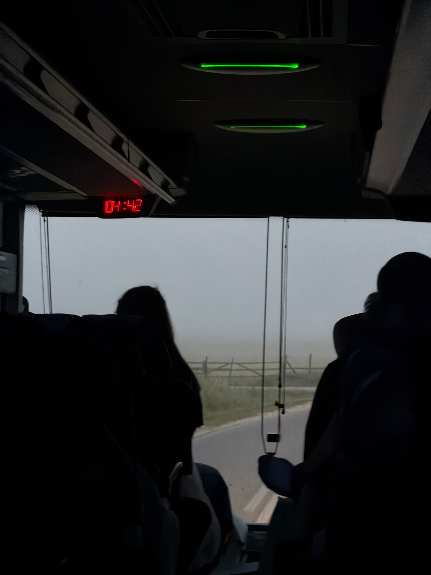 The image, taken inside a vehicle, shows a digital clock displaying "04:42". Two green lights are visible on the ceiling, and the view through the window depicts a foggy landscape with a road and a wooden fence. Silhouettes of passengers are present in the foreground. No specific location in Vidrići, Bosnia and Herzegovina can be identified from the image.