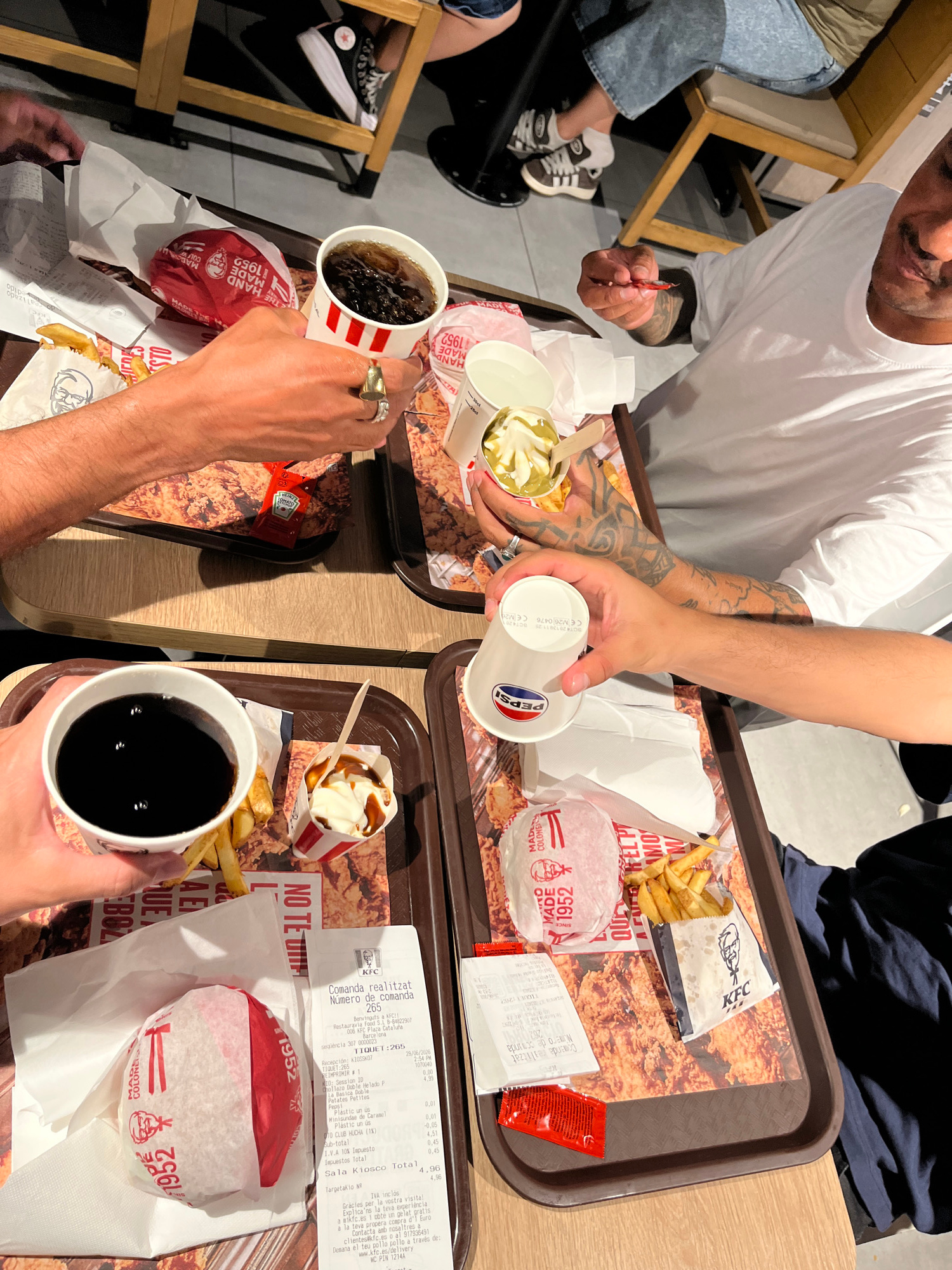 The image shows a top-down view of a KFC meal spread on a table. Several people are present, though only their hands and parts of their bodies are visible, suggesting a group dining together. They are reaching for and holding various food and drink items.
The objects on the table include KFC trays filled with fried chicken and french fries, burgers wrapped in red and white KFC packaging, cups of soda, and containers of ice cream or dessert with caramel topping. There are also receipts and ketchup packets. One person is holding a cup with a Pepsi logo, and another is holding a cup with dark soda. The hands are adorned with rings.
The scene appears to be an indoor setting, likely a KFC restaurant, given the branded packaging and trays. The lighting suggests it could be daytime, but there are no external environmental cues. The activity is clearly people enjoying a meal together, with hands reaching for different items, indicating sharing or distribution of food.
Visible text includes "KFC," "PEPSI," "1952," and various details from the receipts which are in Spanish and Catalan. The receipts mention "Comanda realitzat," "Número de comanda," "Restauravia Food S.L B-848229," "KFC Plaza Cataluña Barcelona," "seqüència," "TIQUET," "Recepción: K10SSK07," dates like "29/06/2026," times, and itemized costs. Phrases like "Gràcies per la vostra visita!" and "Explica'ns la teva experiência a mikfc.es i obté un gelat gratis" are also visible on the receipts, indicating customer feedback incentives and contact information for KFC in Spain. The location context is reinforced by the Spanish and Catalan text on the receipts and the mention of "KFC Plaza Cataluña Barcelona."