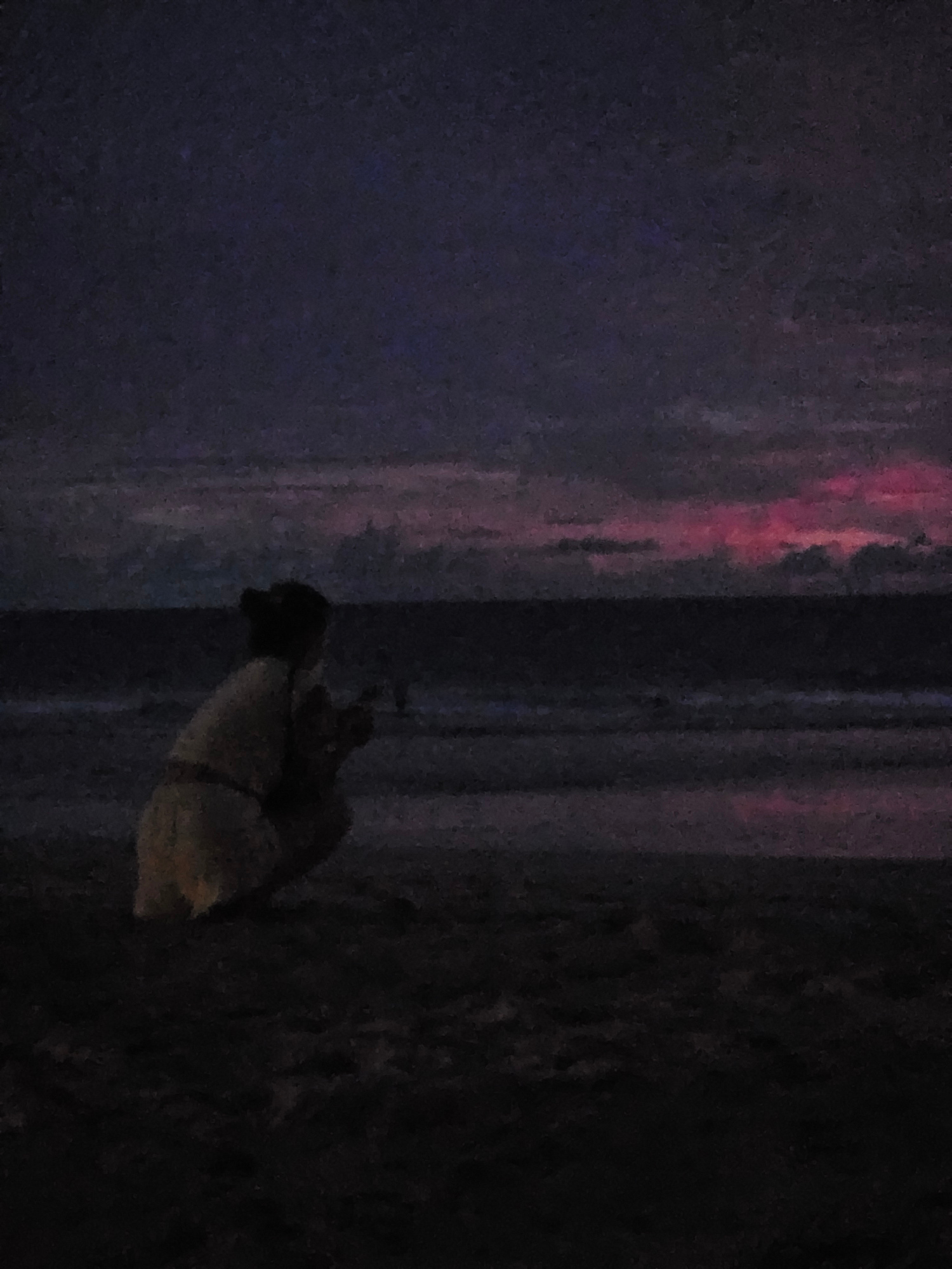 The image shows a close-up, vertically oriented view of a dark, textured surface with streaks of red and pink light. In the foreground, a small, light-colored object with dark accents is visible, possibly an animal or a toy, resting on the surface. The lighting suggests a low-light environment, perhaps dusk or night, with artificial colored lights creating an atmospheric effect. The background is predominantly dark, with some vertical lines suggesting a structured environment like a wall or a fence. The texture of the surface appears rough and somewhat uneven. The overall impression is moody and abstract due to the darkness and the vibrant, streaky colors. Without further context, it's difficult to ascertain the exact nature of the object or the specific activity, but the setting appears to be outdoors given the texture and lighting. The location context of Sakhu, Thailand, doesn't provide direct visual cues within the image itself.