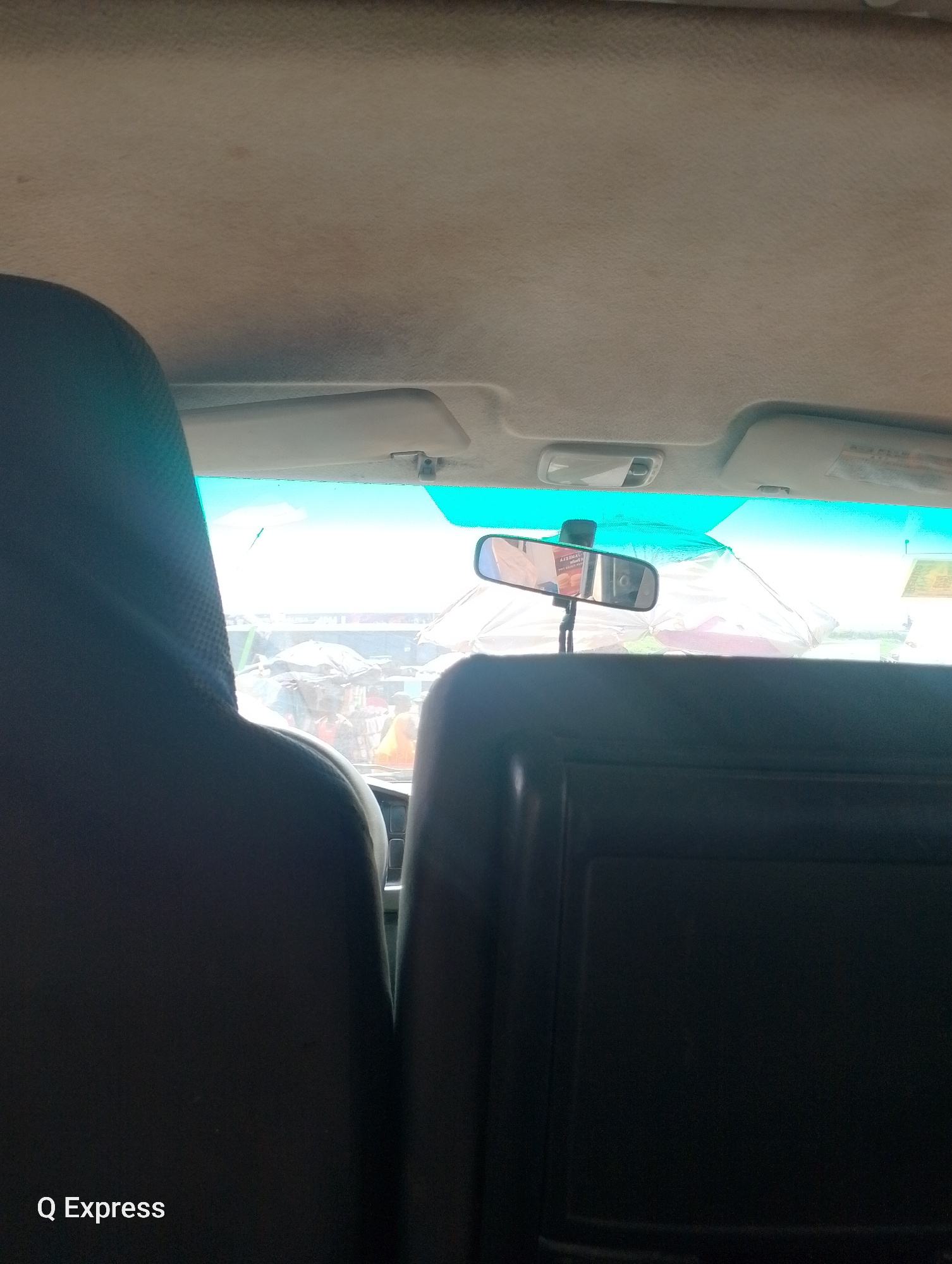 The image is taken from inside a vehicle, looking forward through the windshield. In the foreground, the dark gray headrest of a passenger seat is visible on the left. To the right, a portion of another dark gray seatback is visible. The interior of the vehicle's ceiling, covered in a light beige fabric, takes up the top portion of the image. A sun visor is visible on the right side of the windshield. A rearview mirror is mounted in the center of the windshield, reflecting part of the interior and the exterior scene.
Through the windshield, an outdoor market scene is visible under a bright, clear blue sky. Several individuals are present, some with umbrellas or coverings suggesting stalls or protection from the sun. The market appears to be bustling with activity, though the details of the people and goods are indistinct due to the distance and lighting. The overall impression is of a lively outdoor environment. A logo at the bottom left corner of the image reads "Q Express."