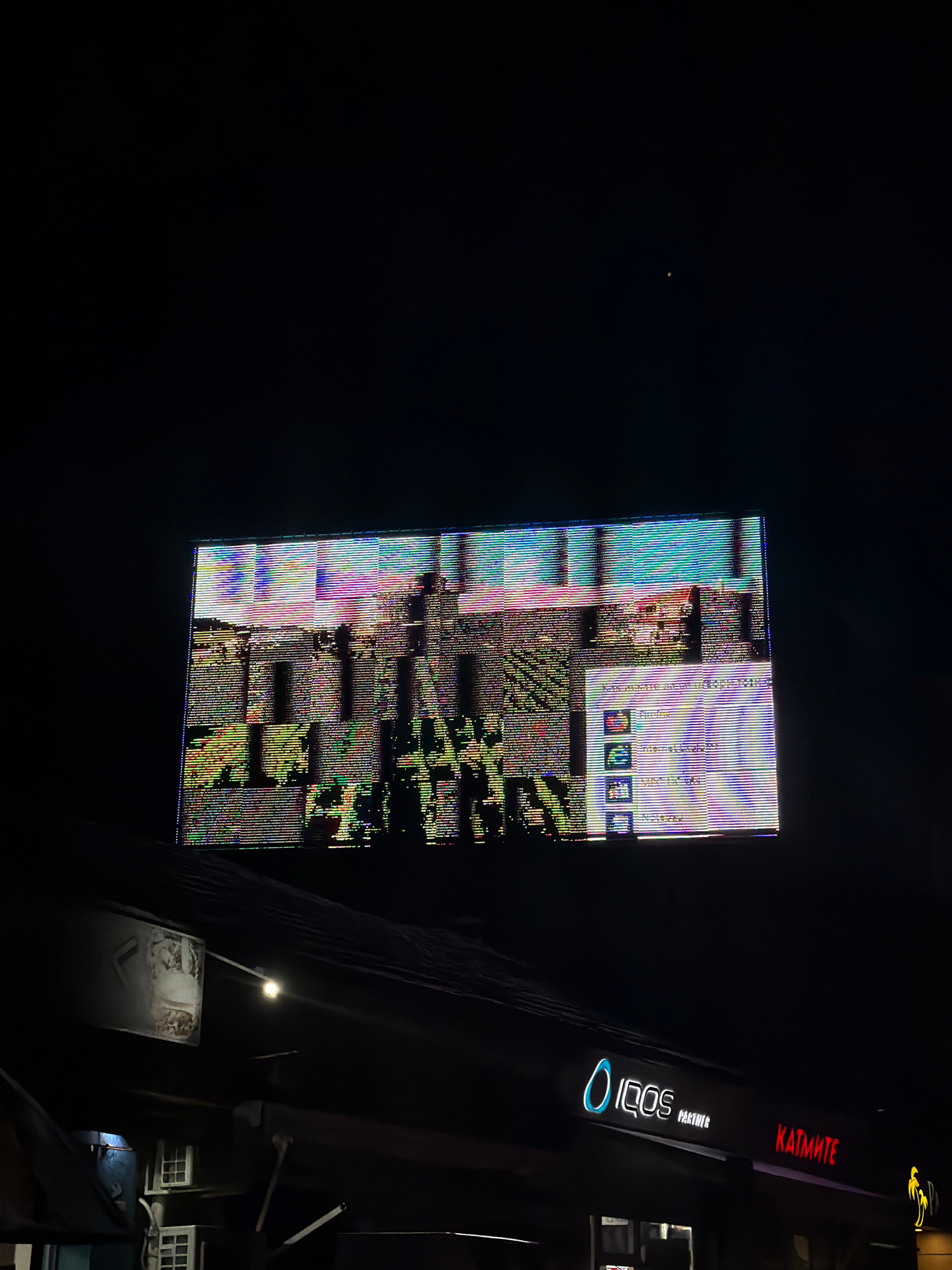 The image shows a damaged LED billboard displaying static and glitching visuals at night in Ruse, Bulgaria. The billboard appears to be situated above a commercial building with illuminated signs for "IQOS PARTNER" and "КАТМИТЕ". The surrounding environment is dark, suggesting it is nighttime. The billboard itself is the primary focus, with its distorted display suggesting a technical malfunction. No people are visible in the image.