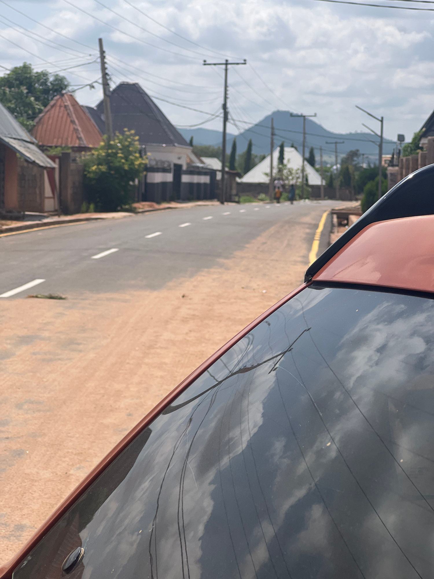 This image shows a view from a vehicle, looking down a paved road in Howayi, Nigeria. The road has a yellow line along the right edge and white dashed lines dividing lanes. The scene is lined with buildings, trees, and utility poles with numerous wires. In the background, hazy mountains are visible under a cloudy sky. The sky reflects on the dark surface of the vehicle in the foreground, showing white clouds. The sun appears to be out, creating a bright, overcast day. The overall impression is a typical street view in a town or village. No people are clearly visible, and no text is present.