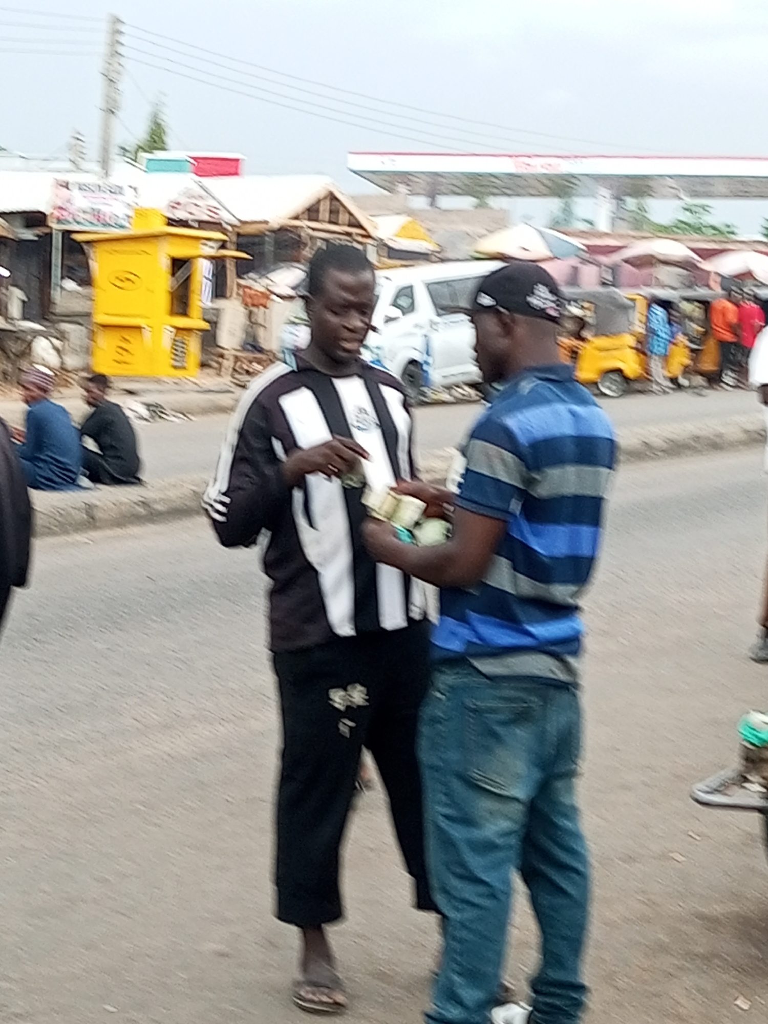 The image depicts a street scene in Gombe, Nigeria, likely in a market or commercial area. Two men are the primary subjects, engaged in an apparent transaction. The man on the left, wearing a black and white striped jacket and black pants, is holding several banknotes. The man on the right, dressed in a blue and grey striped polo shirt and blue jeans, appears to be receiving or examining the money.
In the background, there are various structures, including stalls with corrugated metal roofs and a bright yellow kiosk. A white van is parked nearby, and several yellow and red tricycles (popularly known as "keke napep" in Nigeria) are visible, suggesting a busy transportation hub or a place where goods are exchanged. A white and red structure, possibly a gas station canopy, is visible in the far background. Other individuals are present in the background, some seated and others standing, indicating the presence of a crowd or a bustling marketplace. The overall environment appears to be an open-air setting on a paved road.
