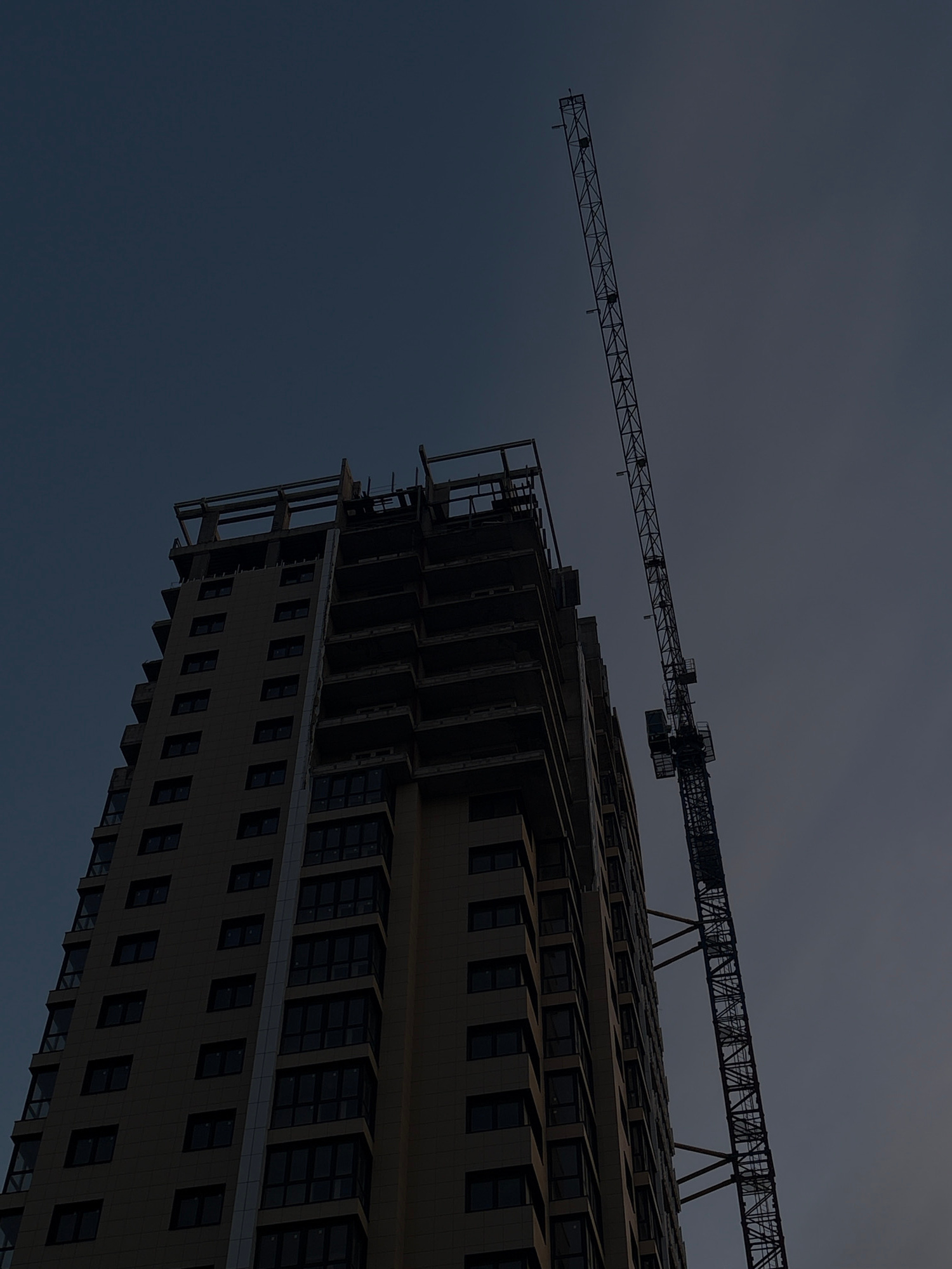 The image shows a tall, unfinished building under construction, with a construction crane positioned next to it. The building is rectangular in shape and appears to be made of concrete and glass. The crane is a large, lattice-boom tower crane, also appearing to be made of steel. The sky in the background is a dark, overcast blue, suggesting either early morning, late evening, or a cloudy day. The overall scene depicts an urban construction site. Without additional context or identifiable landmarks, it is impossible to definitively place this image within the city of Krasnodar, Russia, or any specific location.