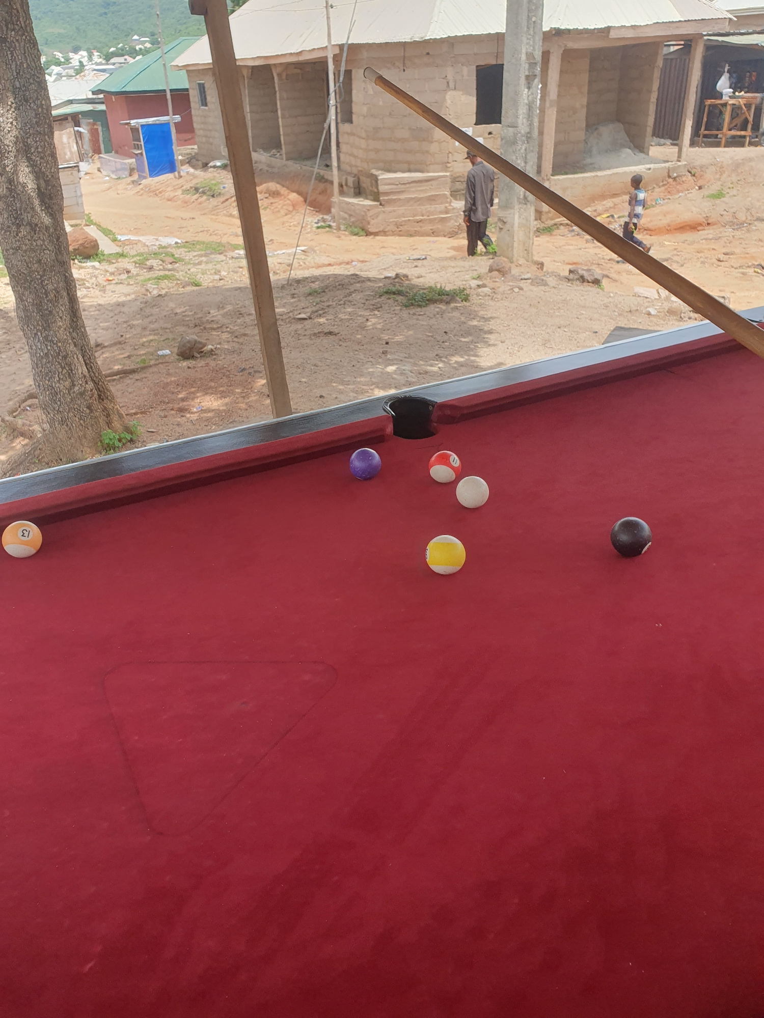 A close-up shot of a red pool table is in the foreground, with several billiard balls scattered across its surface. One ball, numbered 13 and colored orange and white, is positioned near the left edge of the table. A purple ball sits further up on the table. In the mid-ground, a group of billiard balls includes a white ball, a red and white striped ball, and a yellow and white striped ball. A solid black ball is near the right side of the table. In the background, two individuals are visible walking in a dusty, outdoor environment characterized by unfinished brick buildings, a large tree trunk on the left, and sparse vegetation. The scene appears to be in Lokoja, Nigeria, indicated by the architectural style and the general ambiance of the environment.