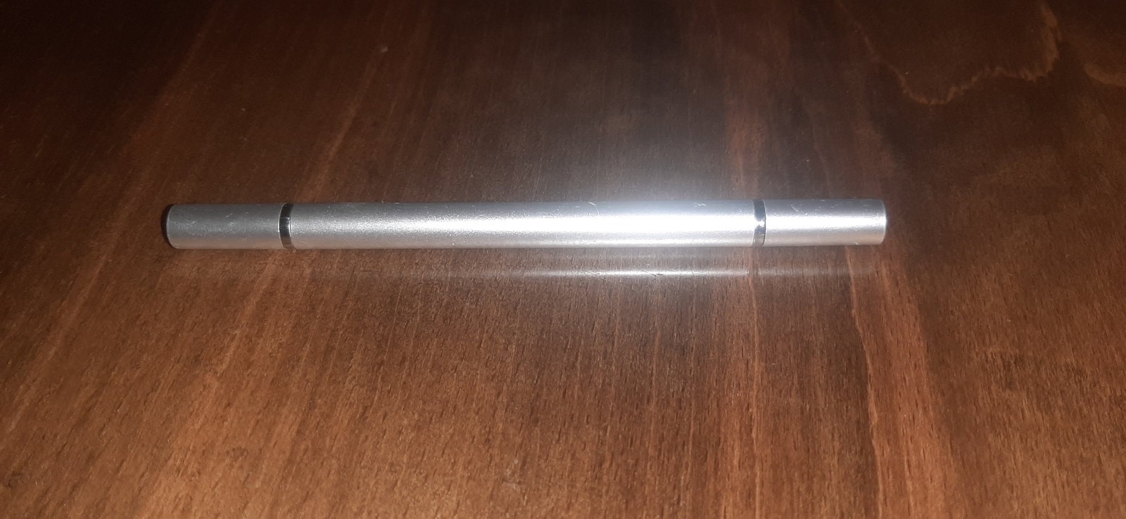 A silver, cylindrical object is centrally placed on a dark wooden surface. The object appears to be a stylus or a pen, with a metallic finish. It is oriented horizontally and reflects the ambient light, creating a bright sheen along its surface. The wooden surface beneath has a visible grain pattern, suggesting a table or countertop. There are no people or other objects visible in the frame. The lighting suggests an indoor setting, possibly with artificial light, as there are no strong shadows indicating natural sunlight. The image is a straightforward product shot, focusing solely on the silver object. The overall mood is neutral and uncluttered. No text is visible on the object or the surface. The context of Yessentukskaya, Russia, provides no visual cues within the image itself.