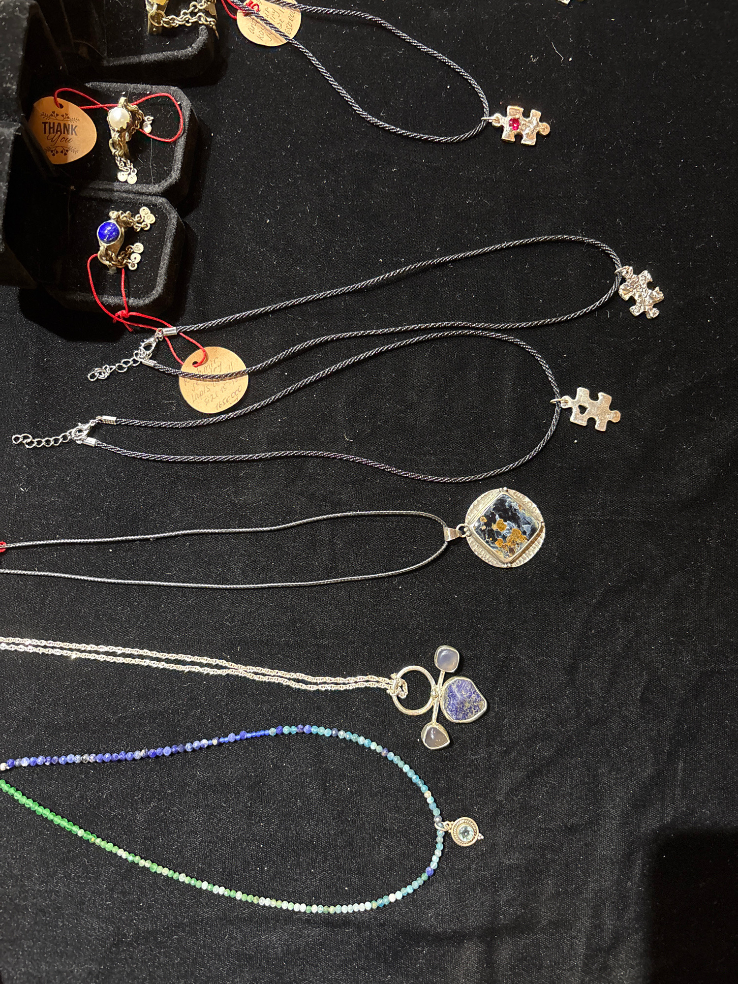 The image shows a collection of necklaces displayed on a black velvet surface. The necklaces vary in design, with some featuring pendants and others made of beads. In the upper left corner, there are two small black velvet jewelry boxes, one containing a ring with a pearl and another with a ring featuring a blue stone. One box has a "THANK YOU" tag. There are also several tags with handwritten text scattered among the necklaces. The scene appears to be a display of jewelry, possibly at a market or a store. The location is given as Unknown, Unknown.