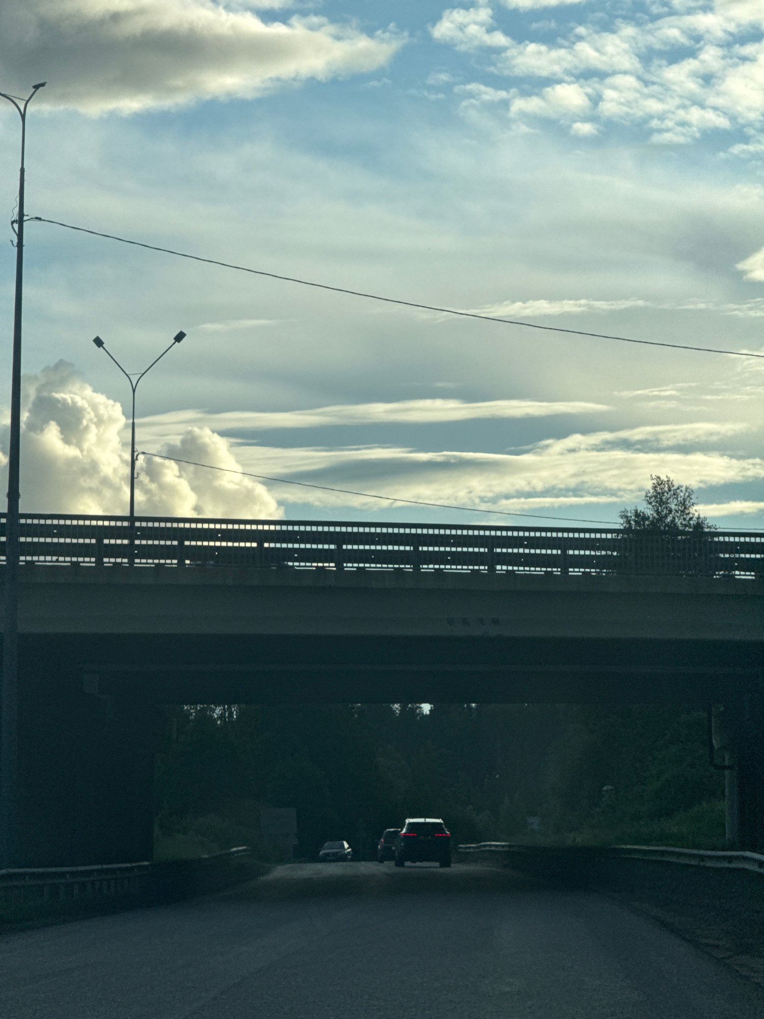 The image shows a view from a road passing under an elevated overpass. The overpass has a metal railing and is supported by a dark concrete structure. Two streetlights are visible on poles, one taller and closer to the left foreground, and another with a Y-shaped support positioned on the overpass. The sky is filled with dramatic clouds, ranging from bright white cumulus to layered, flatter formations, with patches of blue sky visible. A power line stretches across the sky. On the road beneath the overpass, several vehicles are seen. In the middle distance, a white SUV is driving away from the camera, with its brake lights illuminated. To the left of the SUV, a car is partially visible, also moving away. The surroundings beyond the road under the overpass are densely forested. The lighting suggests it is either early morning or late afternoon, with diffused sunlight.