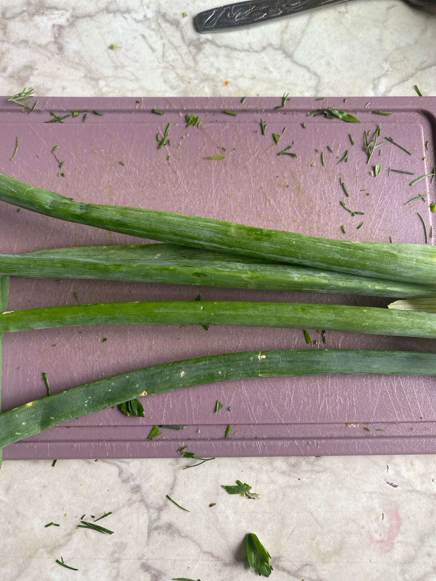 The image shows a close-up, overhead view of several green onions on a light purple cutting board. The cutting board is resting on a marble-patterned surface. Chopped green herbs and some small pieces of green onion are scattered on both the cutting board and the marble surface. A piece of silverware is visible in the upper right corner of the image, partially out of focus. The scene appears to be an indoor kitchen setting, likely in Krupki, Belarus, given the provided context. There are no people visible in the image.