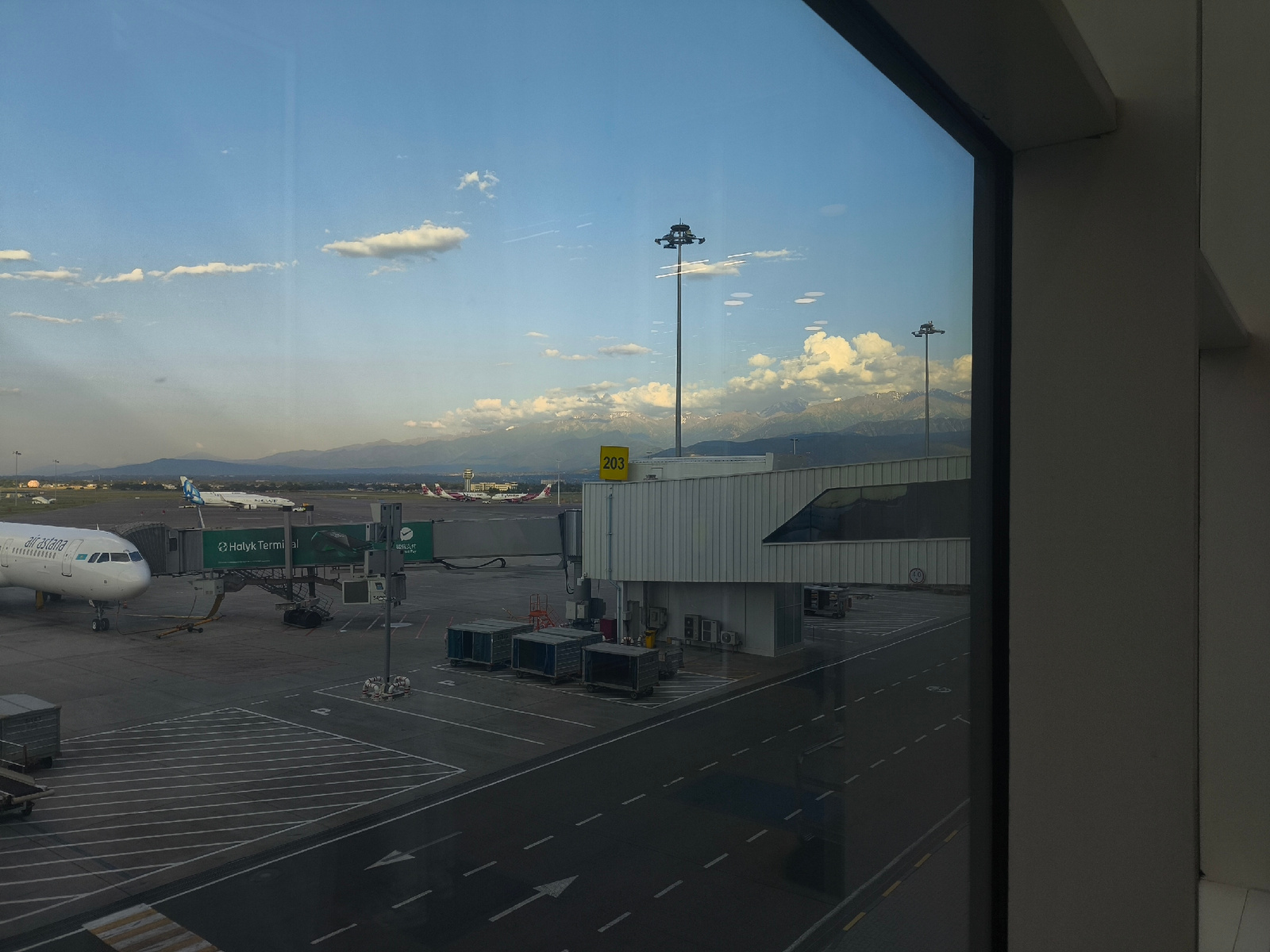 This is an exterior shot of an airport terminal in Almaty, Kazakhstan, viewed through a window. In the foreground, to the left, a white passenger airplane with "airastana" written on its side is parked at a gate, connected by a jet bridge labeled "Halyk Terminal." Several cargo containers are visible on the tarmac, along with markings on the ground. In the background, several other aircraft are parked at gates, and a larger terminal building can be seen. Beyond the airport, a range of snow-capped mountains dominates the horizon under a blue sky with scattered clouds. A tall light pole with multiple lights is prominently featured. The overall scene depicts a typical airport environment with a backdrop of natural scenery.