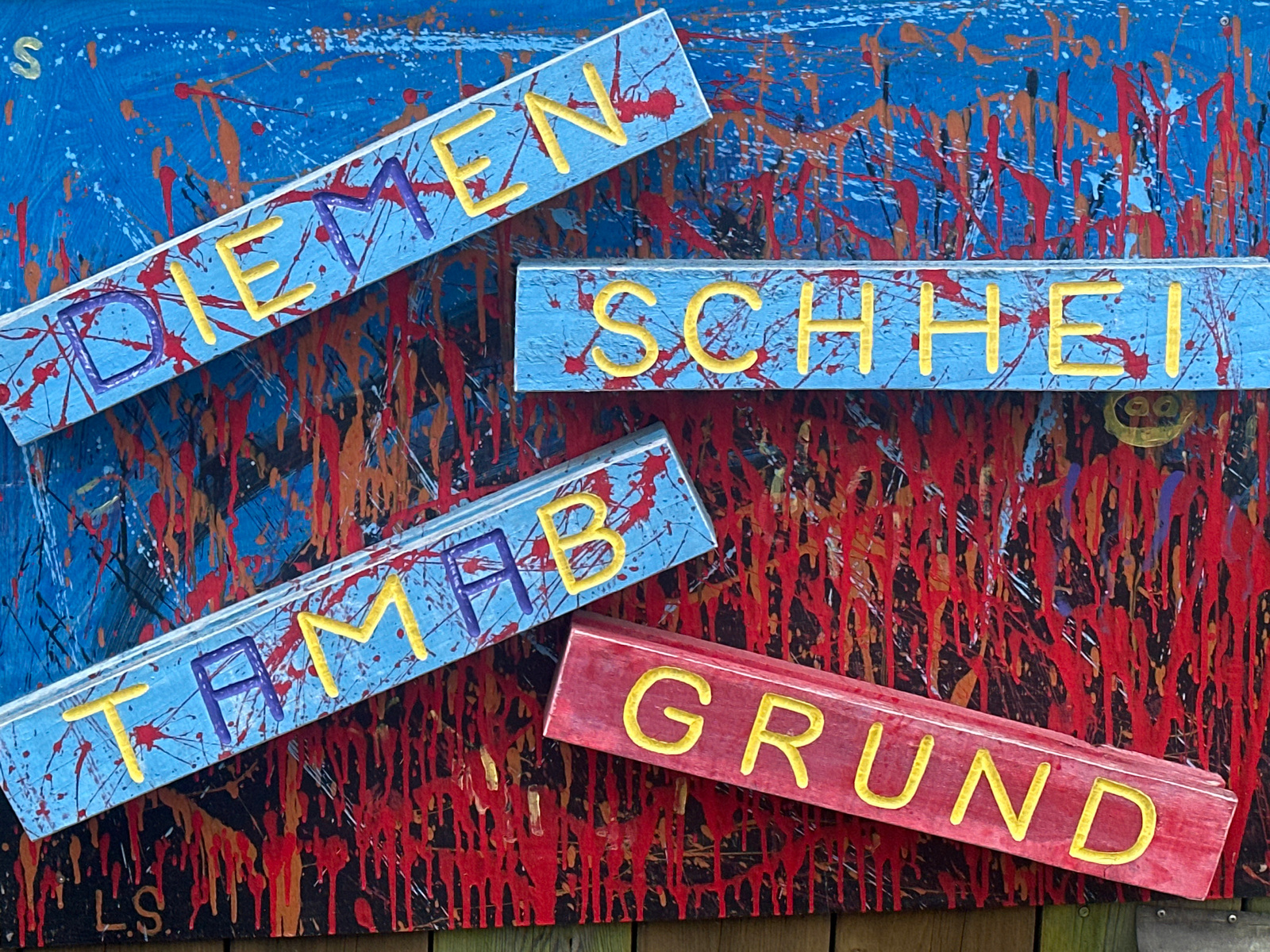 The image displays a piece of abstract art with several wooden planks attached, each bearing painted text. The background is a canvas with a vibrant blue base, splattered with red and orange paint, creating a chaotic and energetic effect.
Attached to the canvas are four wooden planks:
* **Top Left:** A light blue plank with the word "DIEMEN" painted in yellow letters. The letters "DIEM" are outlined in purple.
* **Top Right:** A light blue plank with the word "SCHHEI" painted in yellow letters.
* **Middle Left:** A light blue plank with the word "TAMAB" painted in yellow letters. The letters "TAM" are outlined in purple.
* **Bottom Right:** A red plank with the word "GRUND" painted in yellow letters.
The planks are positioned at various angles, overlapping and creating a layered composition. The overall aesthetic is bold and expressive, with a strong emphasis on color and texture. The artwork appears to be mounted outdoors, possibly as part of a fence or wall, with hints of wooden fence posts visible at the very bottom of the frame. The context of "Unknown, Unknown" suggests this artwork is not tied to a specific, identifiable location.