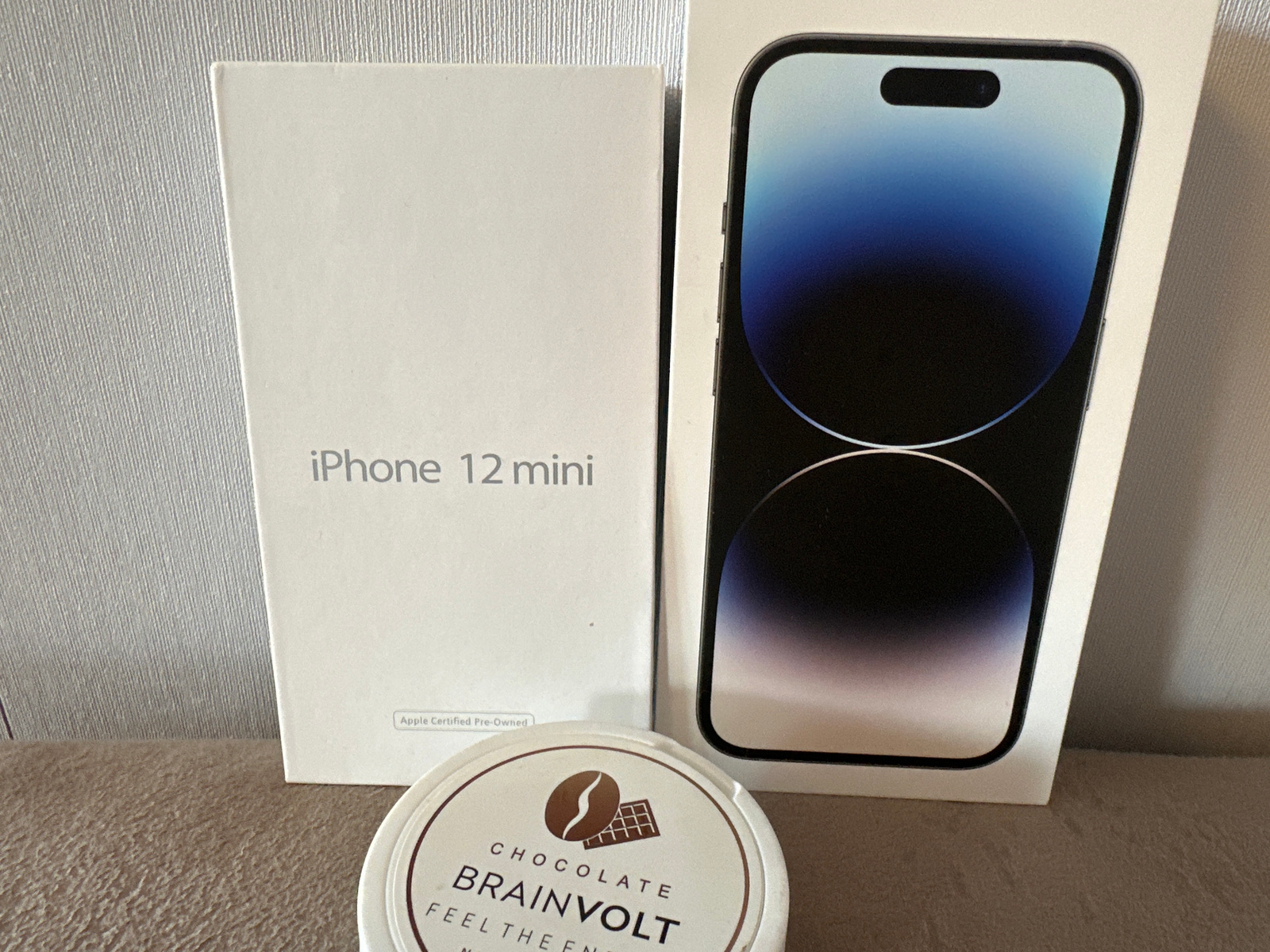 The image shows two iPhone boxes and a small, round container of "BRAINVOLT CHOCOLATE" placed on a textured surface, possibly a table or countertop. The iPhone boxes are white. The larger box on the left displays a portion of a smartphone screen with a dark blue to light blue gradient and a black notch at the bottom. The smaller box on the right has text that appears to be in Russian or a Cyrillic-based language, with the words "iPhone 12 mini" visible. There is also a small label on this box that reads "Apple Certified Pre-Owned." The container of chocolate has text printed in a circular pattern, with "BRAINVOLT" prominently displayed. The lighting suggests an indoor setting, with no specific time of day or weather cues. The overall scene is one of product display, possibly for sale or storage.