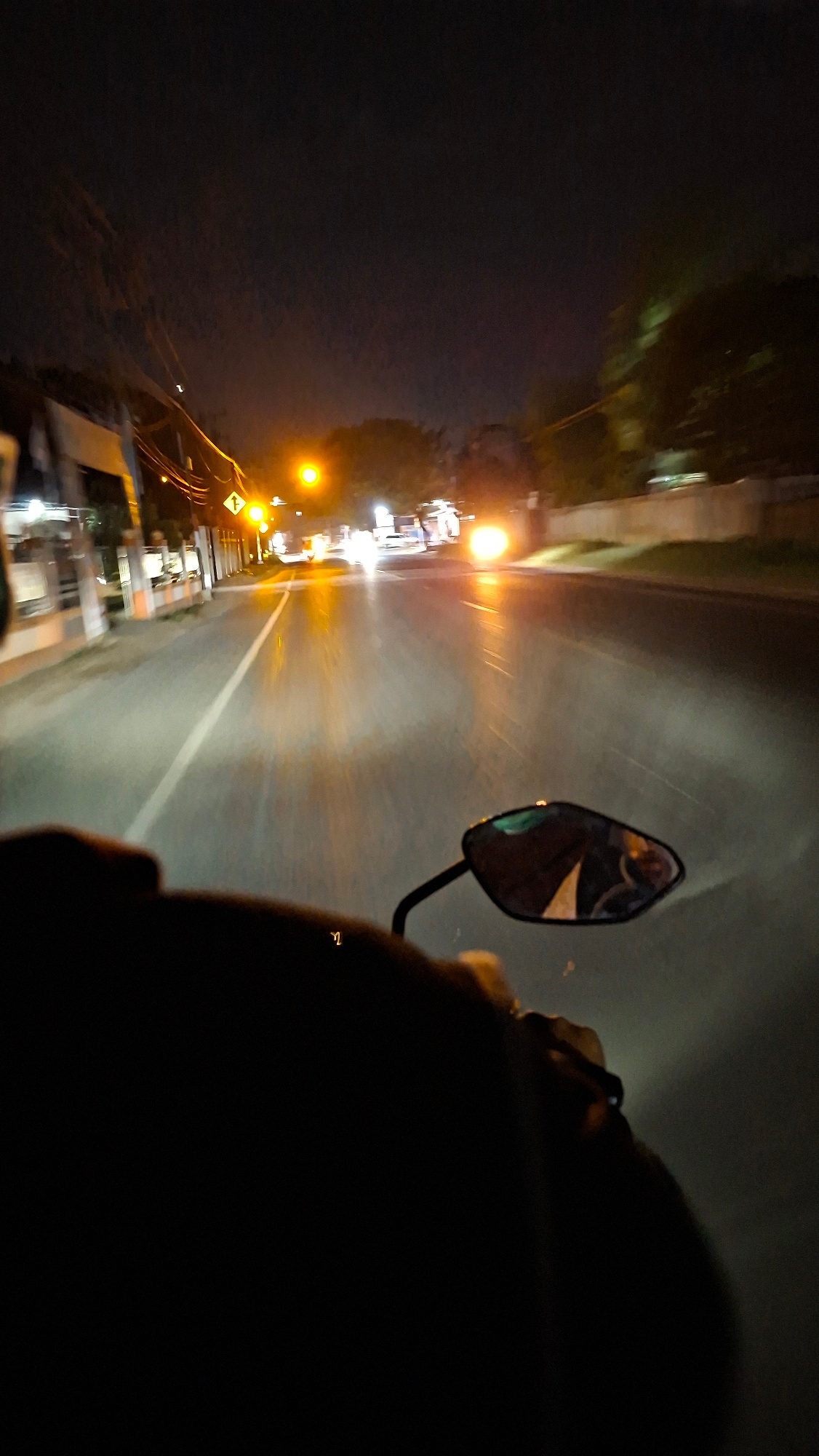 The media file shows a first-person perspective of someone riding a motorcycle at night through Keudebayu, Indonesia. The scene is dimly lit, with streetlights and the headlights of the motorcycle illuminating the road and the surrounding area. The road appears to be paved and has a white line marking its center. On the left side of the road, there are some buildings and trees, with lights from the buildings reflecting on the dark surface. The sky is dark, suggesting it is nighttime.
The rider's shoulder and helmet are visible in the foreground, creating a sense of being in motion. A motorcycle's side mirror is also visible, reflecting a distorted image of the rider and the environment. The rider is likely traveling through a populated area, given the presence of streetlights and buildings. The overall mood is one of travel and exploration in an urban or semi-urban environment at night.
There is a directional sign on the left side of the road which appears to be a yellow arrow indicating a turn. No other text is clearly legible in the media. The time of day is clearly night, and there are no strong indicators of specific weather conditions, though the darkness might suggest a clear night. The activity is riding a motorcycle. The setting is a road in Keudebayu, Indonesia.