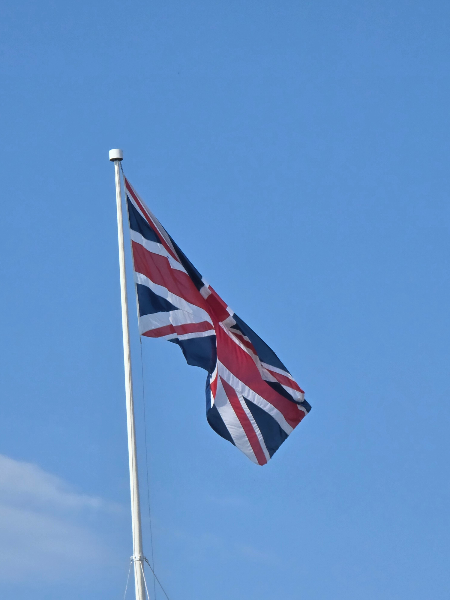 A Union Jack flag, featuring its characteristic red, white, and blue design, is prominently displayed against a clear blue sky. The flag is attached to a tall, white flagpole, which extends vertically through the frame. The flag is caught by the wind, causing it to billow outwards, revealing its intricate pattern. The lower portion of the flagpole is visible, anchored by thin ropes attached to its base. The scene suggests an outdoor setting, likely a public space or monument, in London, United Kingdom, given the prominent display of the Union Jack. There are no people or other objects immediately visible in the frame.
