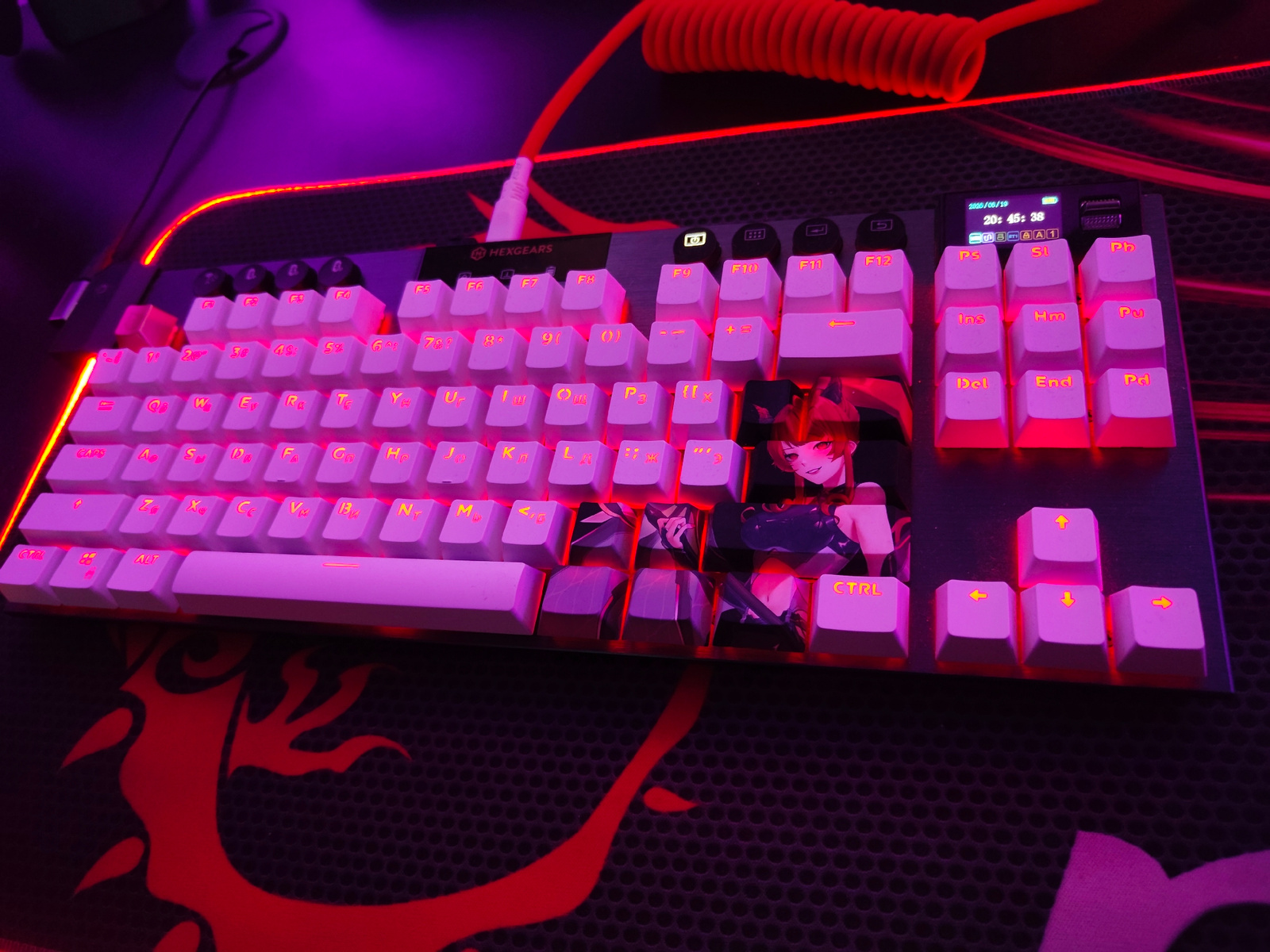 An overhead, slightly angled shot displays a custom mechanical keyboard with white keycaps that are illuminated by pink and red LEDs. The keyboard is positioned on a black mousepad with a red fiery graphic. The keyboard features various key labels, including alphanumeric characters, function keys (F1-F12), and navigational keys. Some of the keycaps, particularly on the right side, are decorated with anime-style characters and artwork.
To the upper left, a black computer mouse is partially visible, connected to a cable that extends towards the keyboard. A coiled red cable, likely for the keyboard, is prominent in the upper right quadrant of the image. The background consists of a dark surface, illuminated by the purple and red glow emanating from the keyboard's lighting, creating a vibrant and atmospheric setting. The overall scene appears to be a desktop setup, likely in a gaming or personal computer environment.
The location specified, Borova, Ukraine, is not discernible from the visual information presented in the image. There are no elements within the photograph that provide any geographical or locational context to Borova, Ukraine.