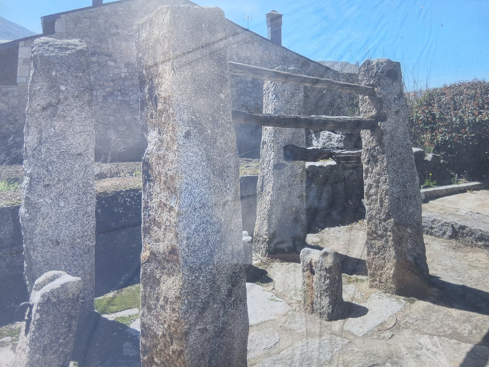 This image depicts a stone structure in Navaluenga, Spain, likely a dolmen or megalithic monument, characterized by several large, weathered granite uprights supporting wooden beams. In the background, a stone building with a tiled roof and a chimney is visible. The surrounding environment includes a stone pathway, dry vegetation, and a dense green shrubbery. The scene is illuminated by sunlight, casting shadows on the ground.