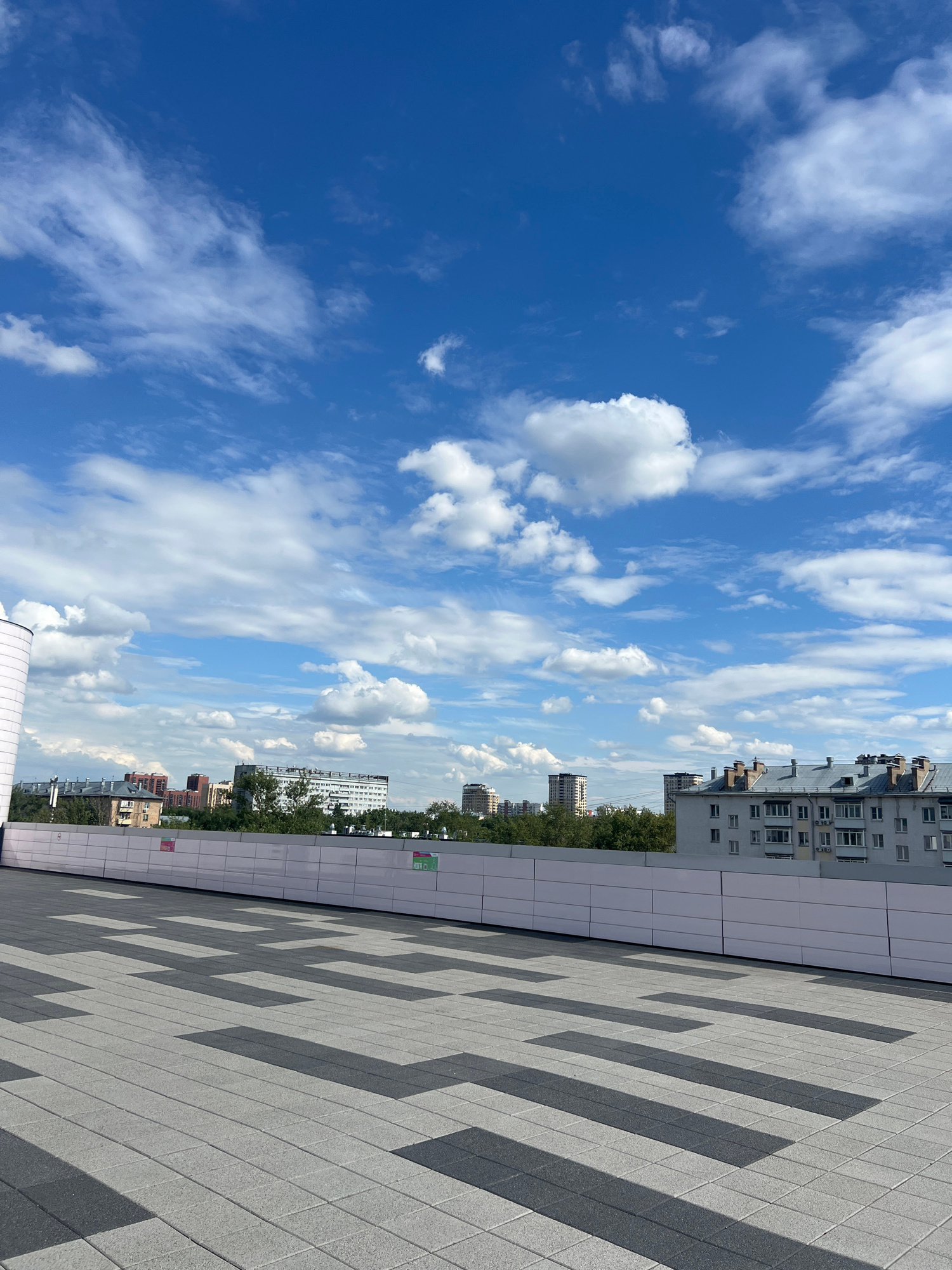 The image displays a modern building facade with a patterned tiled walkway in the foreground. The building is light-colored, with rows of windows visible. Behind the building, a cityscape of other buildings and trees can be seen under a bright blue sky filled with scattered white clouds. The scene appears to be set on a rooftop or elevated walkway, providing a view of the urban environment. The lighting suggests it is daytime, with the sun casting shadows on the tiled ground, indicating a clear day. There are no people visible in the image. Some faint red signage with white text is visible on the building facade in the background, but the text is not legible. The overall mood is serene and the setting is urban.
