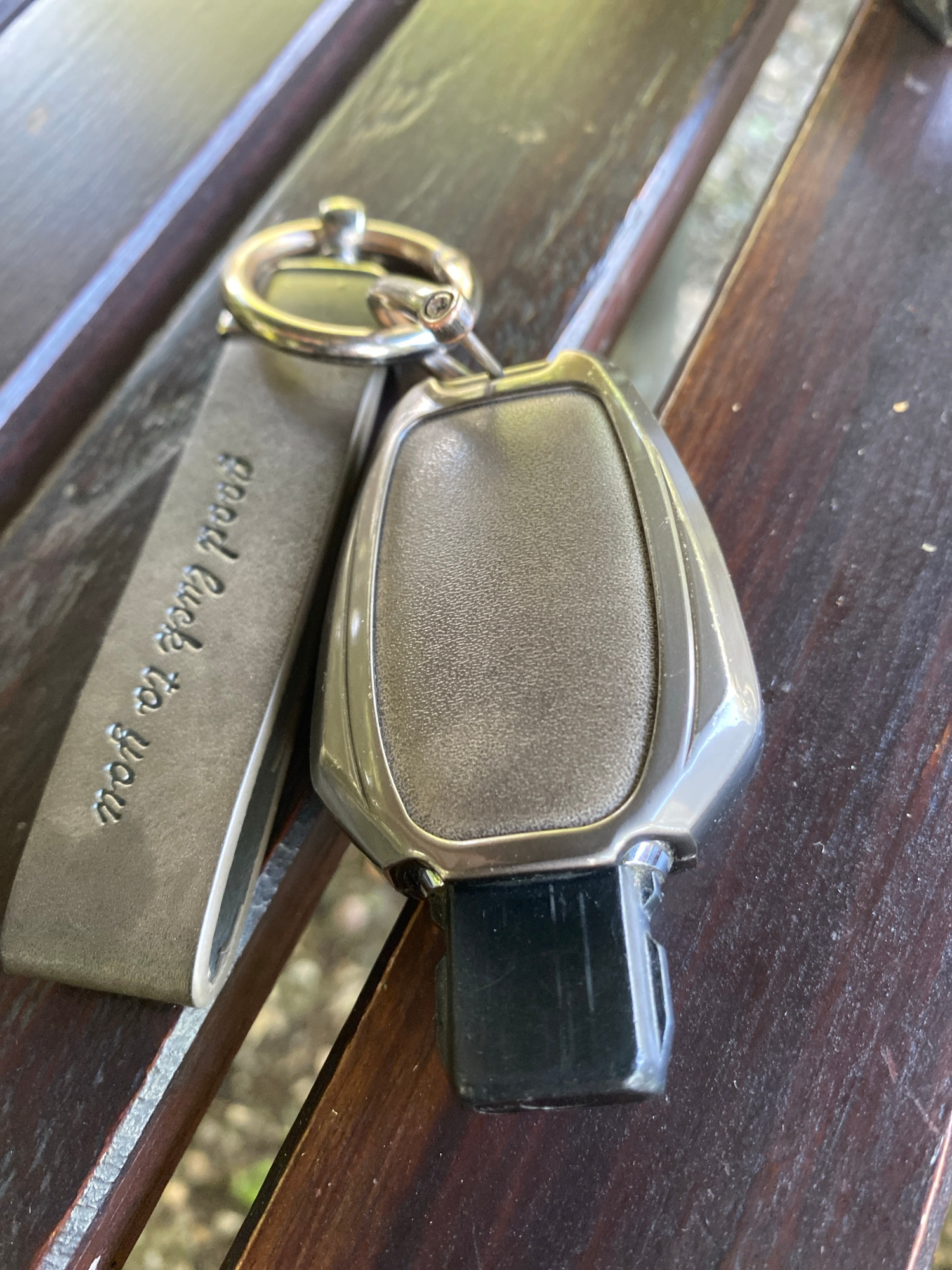 A car key fob with a textured gray insert and a black electronic component rests on a wooden bench. Attached to the key fob by a metal ring is a flat, gray leather strap. Embossed on the leather strap in cursive font are the words "good luck to you". The bench appears to be outdoors, with a blurry background of greenery and dappled sunlight, suggesting a daytime setting with pleasant weather. The overall scene is a simple, still life composition focusing on the keychain. The location context of Varna, Bulgaria, does not provide specific visual cues in the image itself.