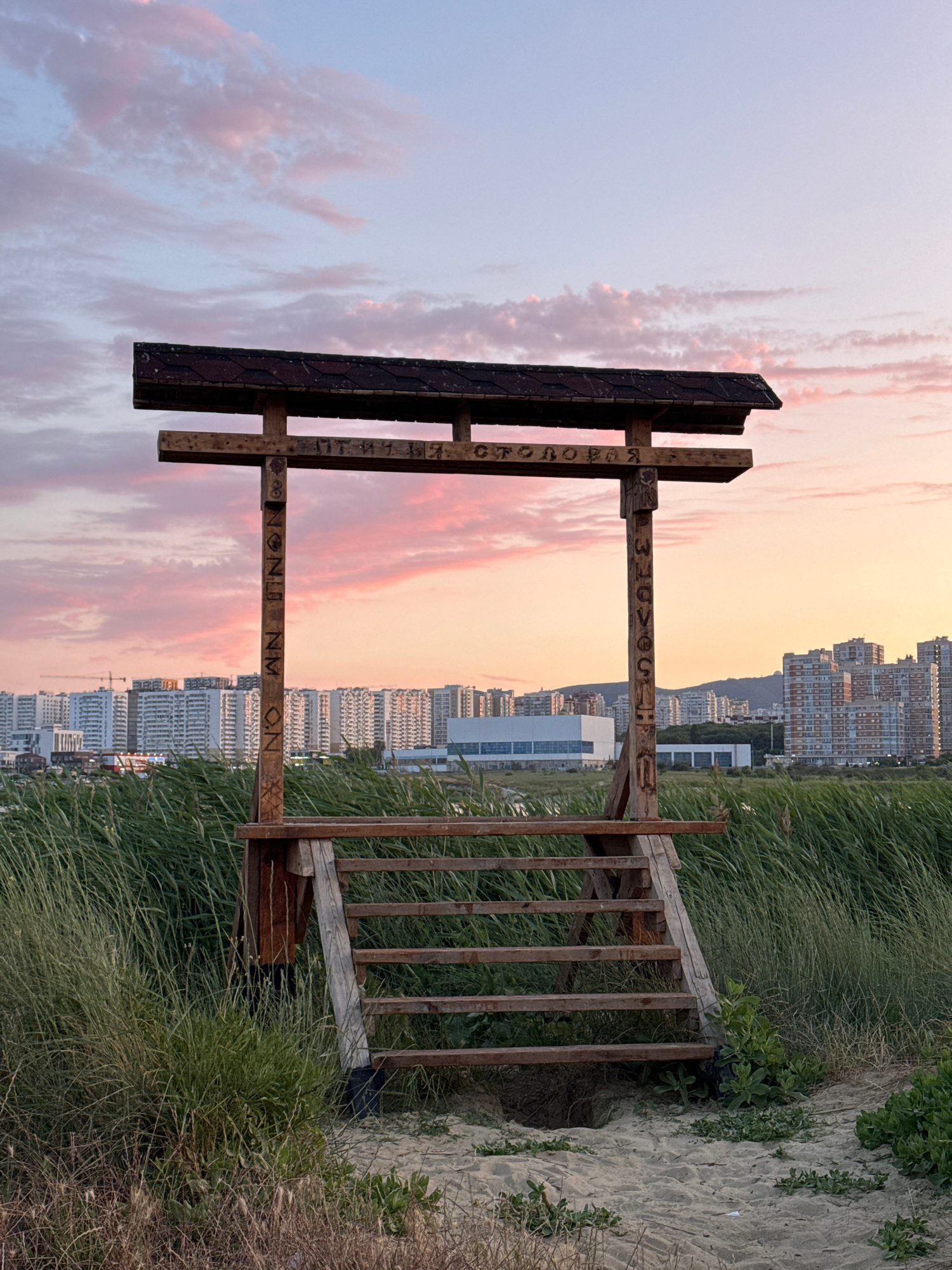 The image shows a wooden structure with a roof, resembling a gateway or small pavilion, situated on a sandy beach. The structure has carved Cyrillic letters on its posts. Behind the structure, a dense field of tall green grass undulates, and beyond that, a cityscape with numerous tall apartment buildings is visible. The sky above is a soft pink and blue, indicating either sunrise or sunset. The overall setting appears to be a coastal area within the city of Novorossiysk, Russia. No people are present in the image.