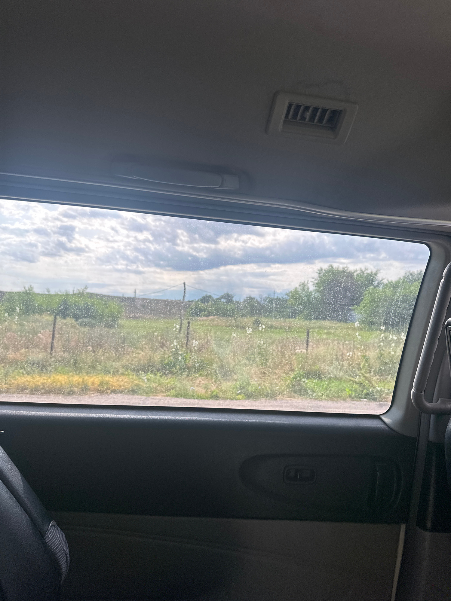 This image, taken from inside a vehicle, shows a view through a window overlooking a rural landscape. The window has visible water droplets or streaks on it. Outside, there is a field with dry grass, some green foliage, and scattered white flowers. A simple wire fence with wooden posts runs horizontally across the middle ground. In the background, there are trees and a cloudy sky.
The interior of the vehicle is visible in the foreground. The headliner of the vehicle's ceiling is grey, with an air vent positioned centrally. The door panel is dark grey or black, with a door handle and a button or latch visible. A portion of a dark seat is also visible on the left side of the frame.
The description does not contain any information about the city of Қасымбек, Kazakhstan, or any people or actions.
