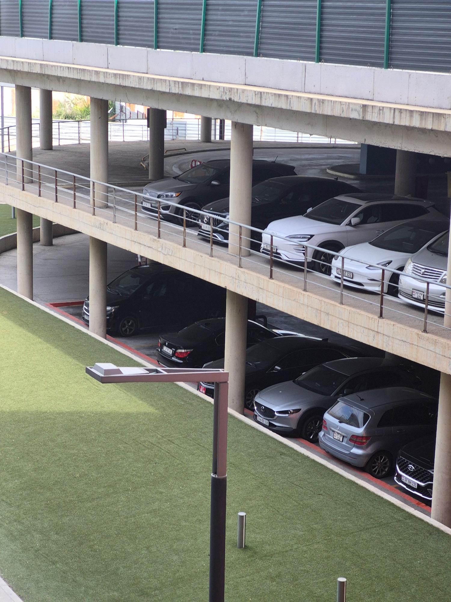 The image captures a multi-level parking structure, likely in Yerevan, Armenia, as suggested by the license plates visible on some of the vehicles. The structure is characterized by concrete pillars and ramps, with a metal barrier along the edges. The lower levels are filled with a variety of parked cars, including several dark-colored sedans and SUVs, and a few lighter-colored vehicles. Artificial turf covers a significant portion of the ground level in the foreground, with a modern-looking lamp post situated on it. The upper levels of the parking structure are visible, showing more concrete and structural elements, with some glimpses of the surrounding urban environment. The scene is devoid of any human presence.