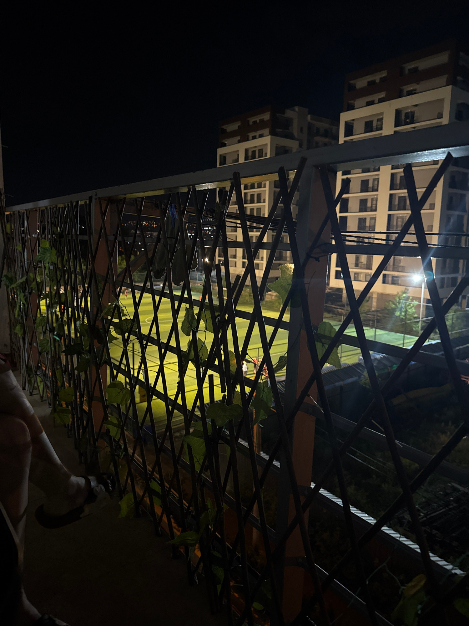 The image depicts a nighttime view from a balcony overlooking a brightly lit sports field, likely a tennis or multi-purpose court, in Tbilisi, Georgia. The foreground features a dark balcony with a decorative lattice fence adorned with artificial green leaves. On the left, a person's legs and feet are visible, with one foot resting on the floor and the other in a sandal. Beyond the fence, the sports field is illuminated by overhead lights, revealing its green surface and visible court lines. Several figures, appearing to be people engaged in an activity on the field, are discernible in the distance. In the background, modern apartment buildings with illuminated windows rise against the dark night sky. The scene captures a typical urban evening setting in Tbilisi, with recreational activity taking place amidst residential architecture.